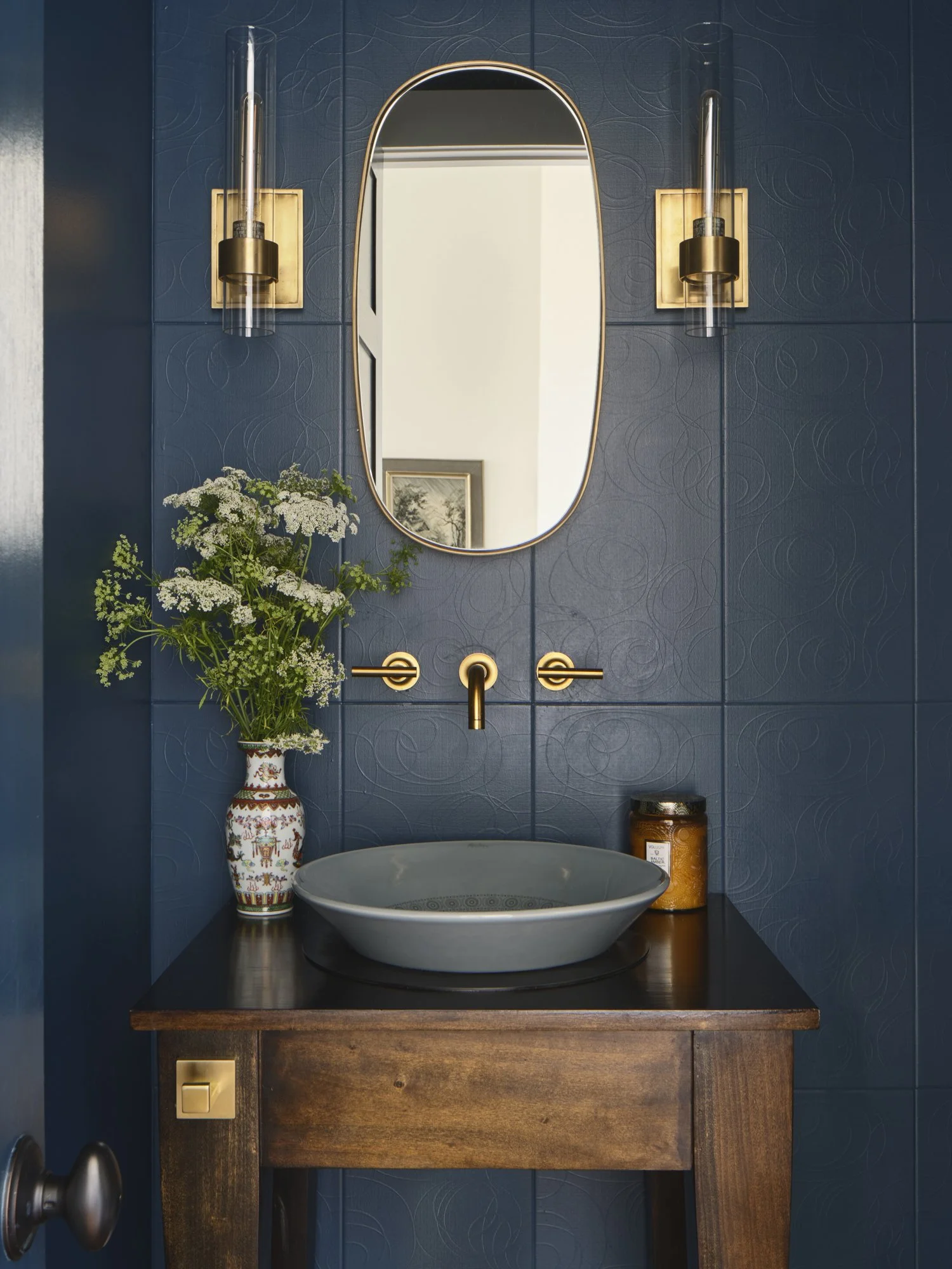A modern bathroom with dark blue textured tiled wall, oval mirror, brass wall sconces, a dark wooden vanity, a grey vessel sink, a vase with flowers, and a candle.
