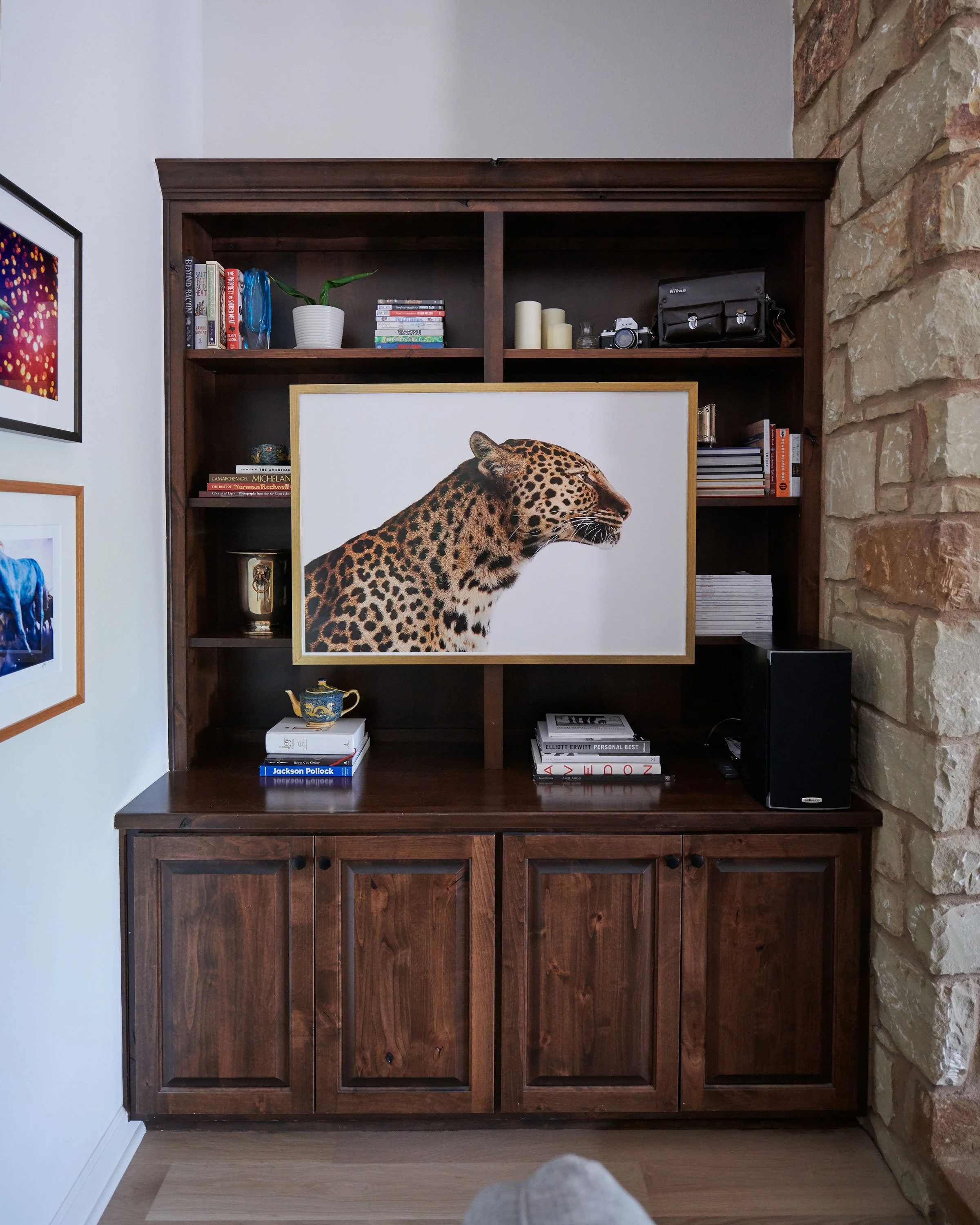 A wooden bookshelf with a photo of a leopard in the middle, surrounded by books, candles, a plant, and a camera, against a brick wall and a white wall with framed art.