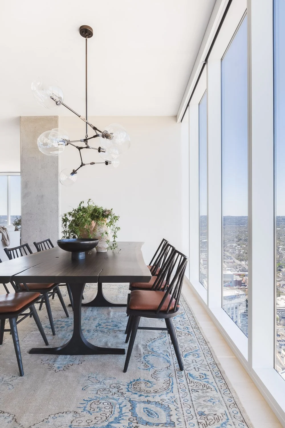 Modern dining room with a large dark wood table, six black chairs with brown seats, a decorative plant in a large black bowl, a contemporary chandelier, and panoramic city views through floor-to-ceiling windows.