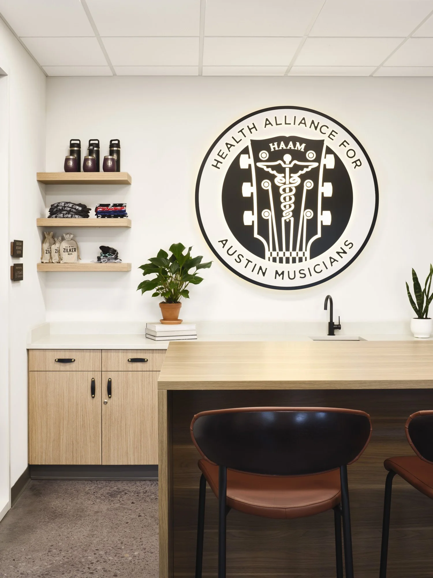Interior of an office or reception area with a large logo on the wall reading 'Health Alliance for Austin Musicians' featuring a guitar with medical symbols, wooden shelves with water bottles and folded clothes, a white counter with a plant and stack of notebooks, and chairs around a wooden counter.