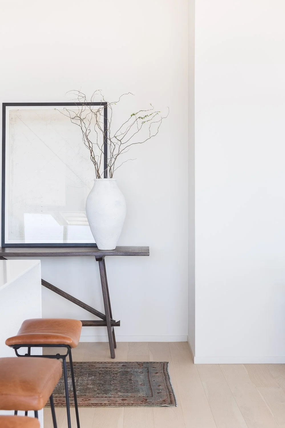 A white vase with bare, twisting branches on a dark wooden table against a white wall with a framed print in the background.