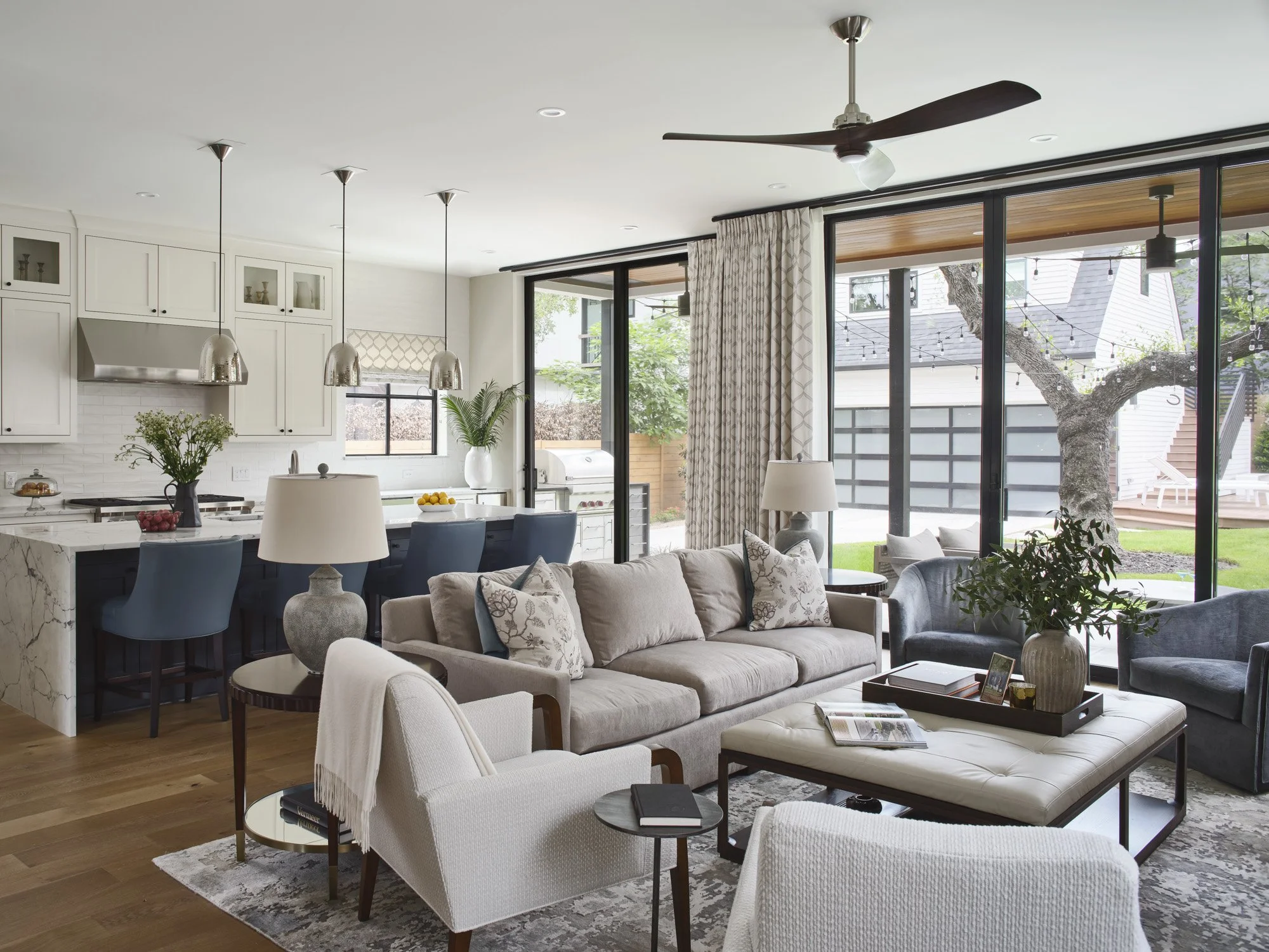 Open-concept living room and kitchen with large windows, gray and beige furniture, and a view of a backyard with a tree and patio lights.