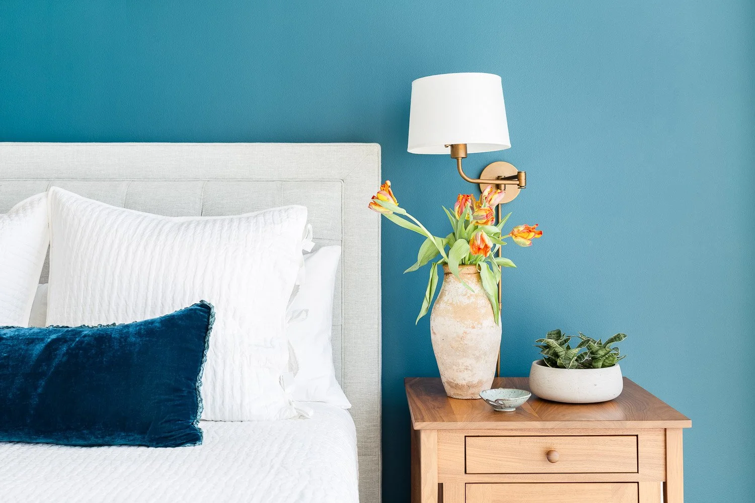 A bedroom with a white headboard, white pillows, a blue velvet pillow, a wooden bedside table, a large ceramic vase with orange and yellow tulips, a small bowl, and a pot with green plants, against a teal wall.