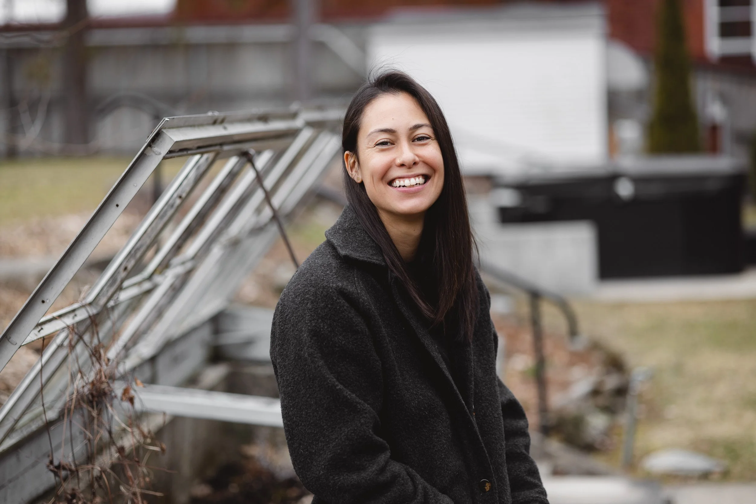 A smiling young woman with long dark hair, wearing a black coat, standing outdoors near a backyard fence and a ladder.