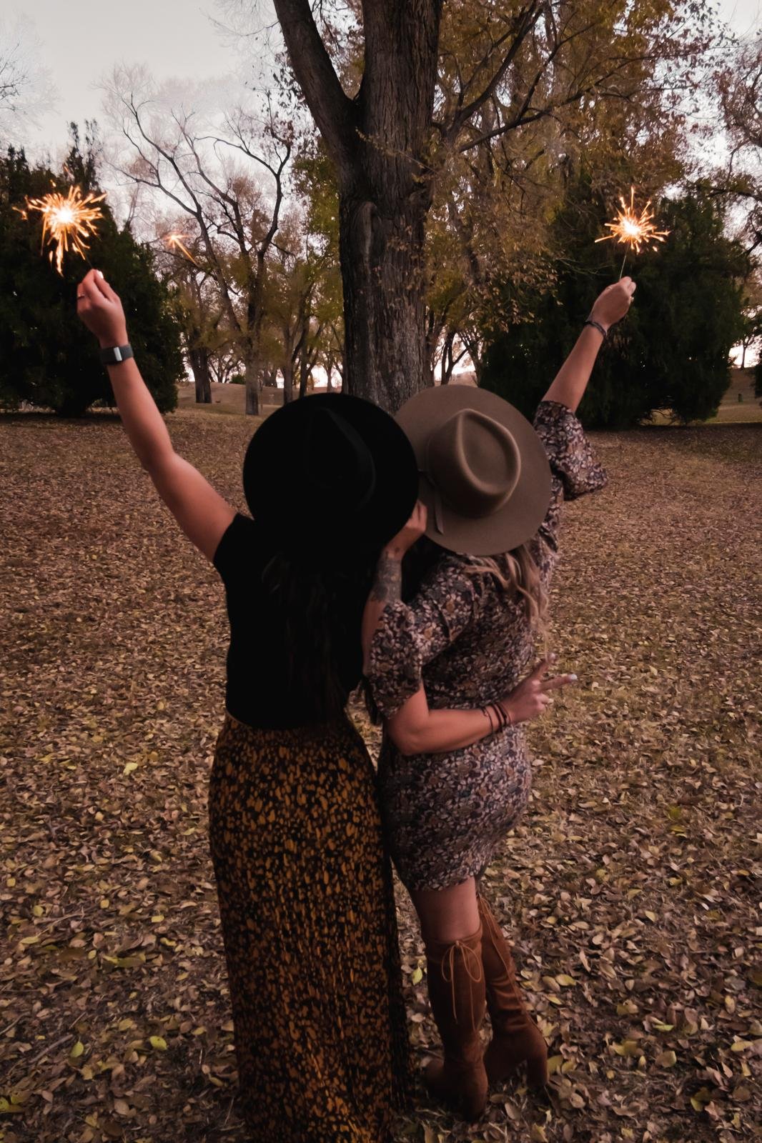 Two women celebrating outdoors at dusk, wearing hats and holding sparklers against a backdrop of trees and fallen leaves.