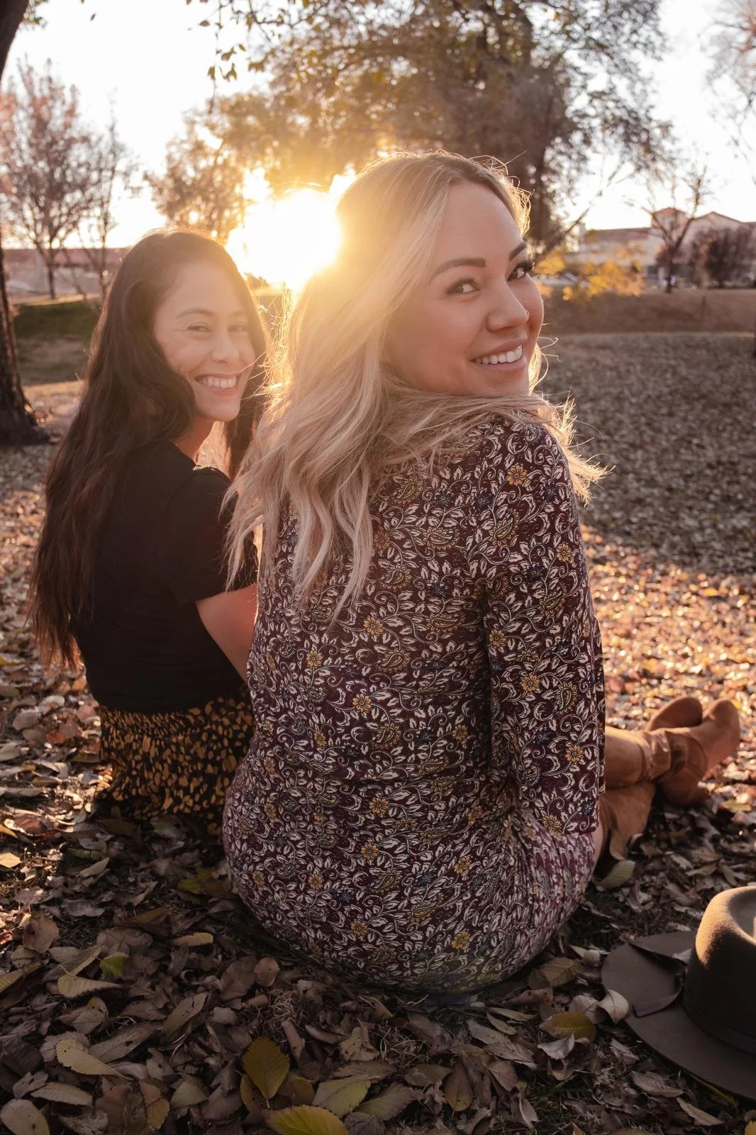 Two women sitting on autumn leaves in a park at sunset, smiling at the camera.