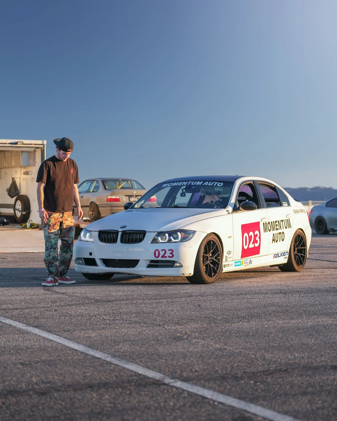 A white race car with black wheels parked in a lot, next to a man in camouflage pants, a black t-shirt, and a cap. The car has racing stickers and the number 023, with a backdrop of other vehicles and mountains in the distance.