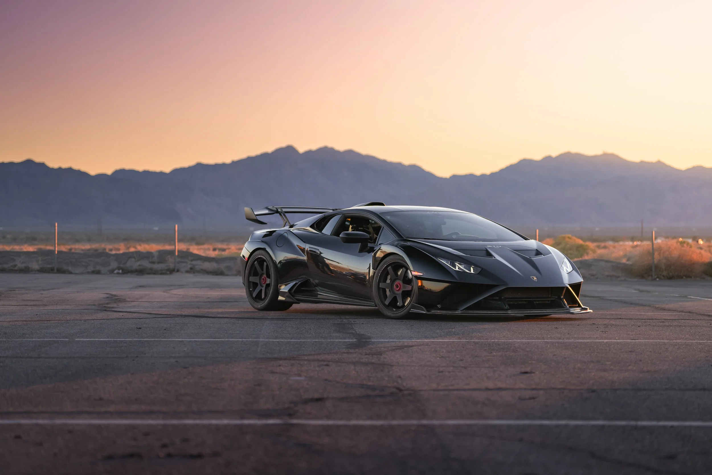 Black Lamborghini sports car parked on an empty desert road during sunset with mountains in the background.