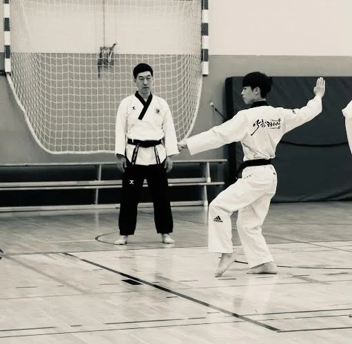 Two martial artists practicing in a gymnasium. One is demonstrating a technique while the other watches.