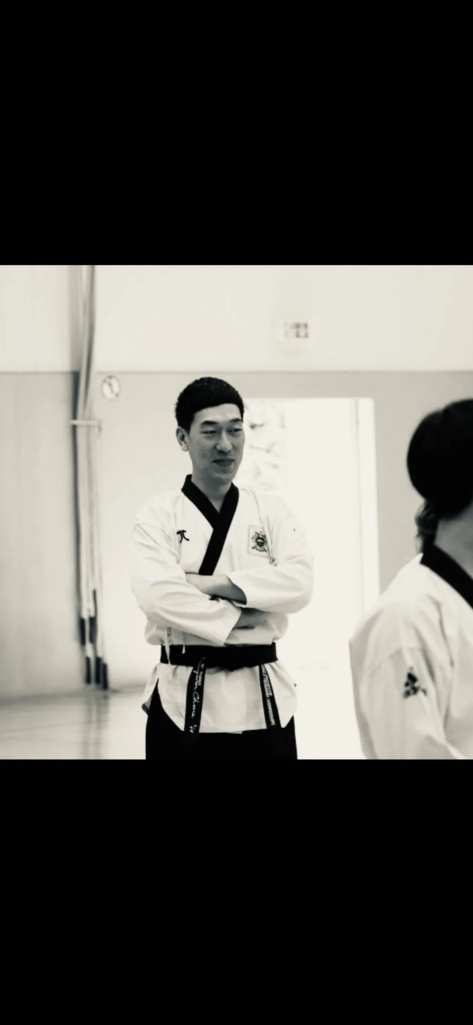 Black and white photo of a martial arts instructor in a taekwondo uniform with arms crossed, standing in a training gym with another student partially visible.
