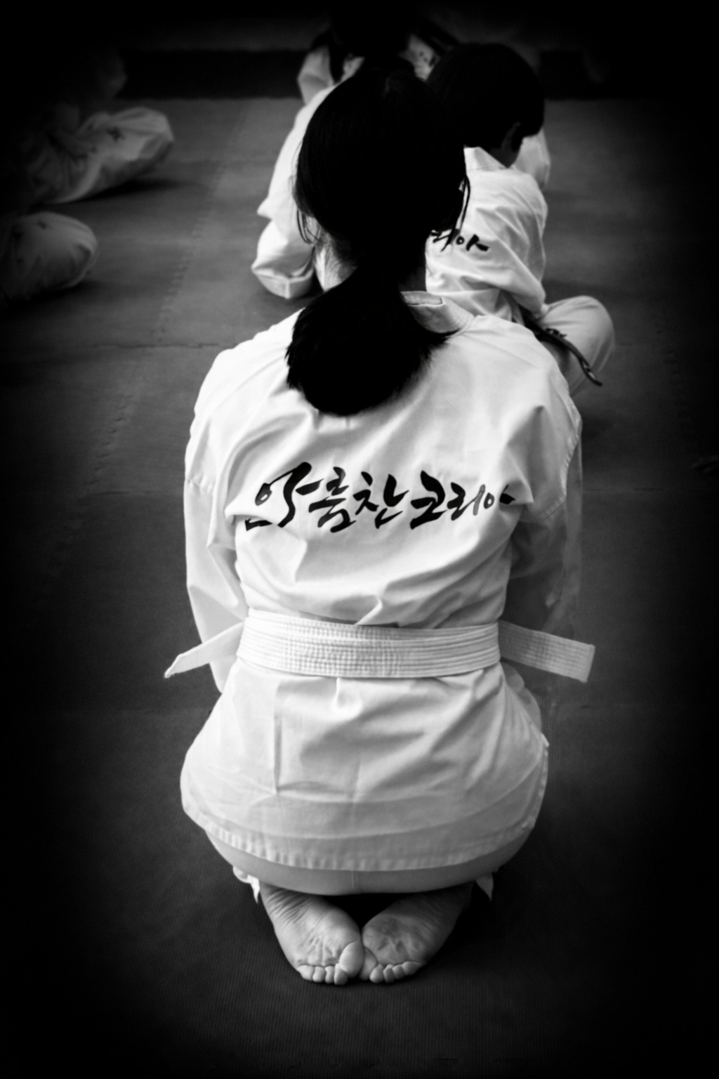 A person kneeling on the floor in a martial arts uniform with Korean writing on the back, practicing martial arts in a dojo with other students.