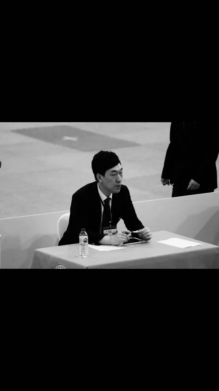 A young man in a suit sitting at a table with a water bottle, a tablet, and some papers, possibly at a conference or event. The image is in black and white.
