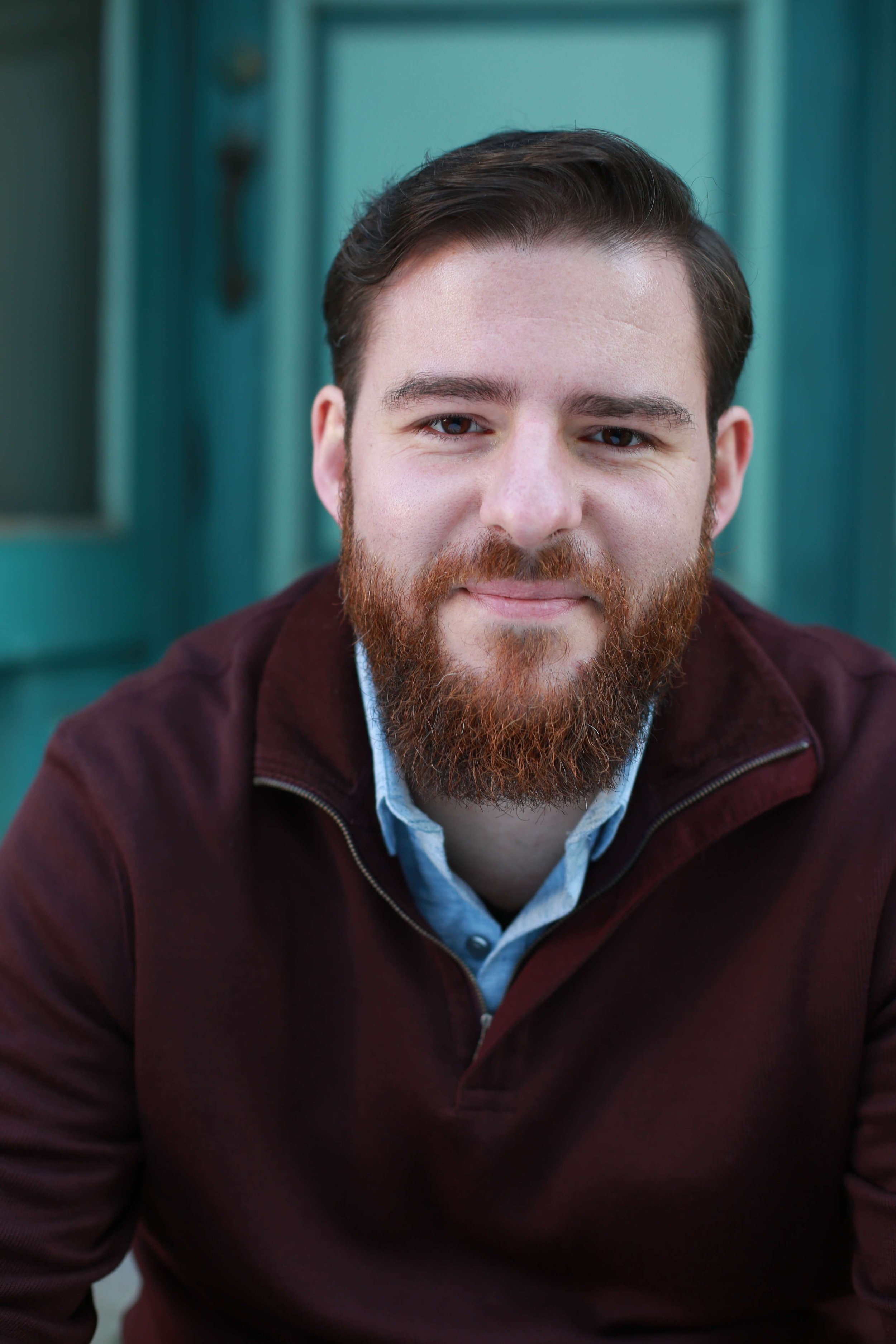 Man with a reddish brown beard and brown hair grins closed-mouth looking at camera, wearing a burgundy quarter zip over a light blue button up against a blurred teal background