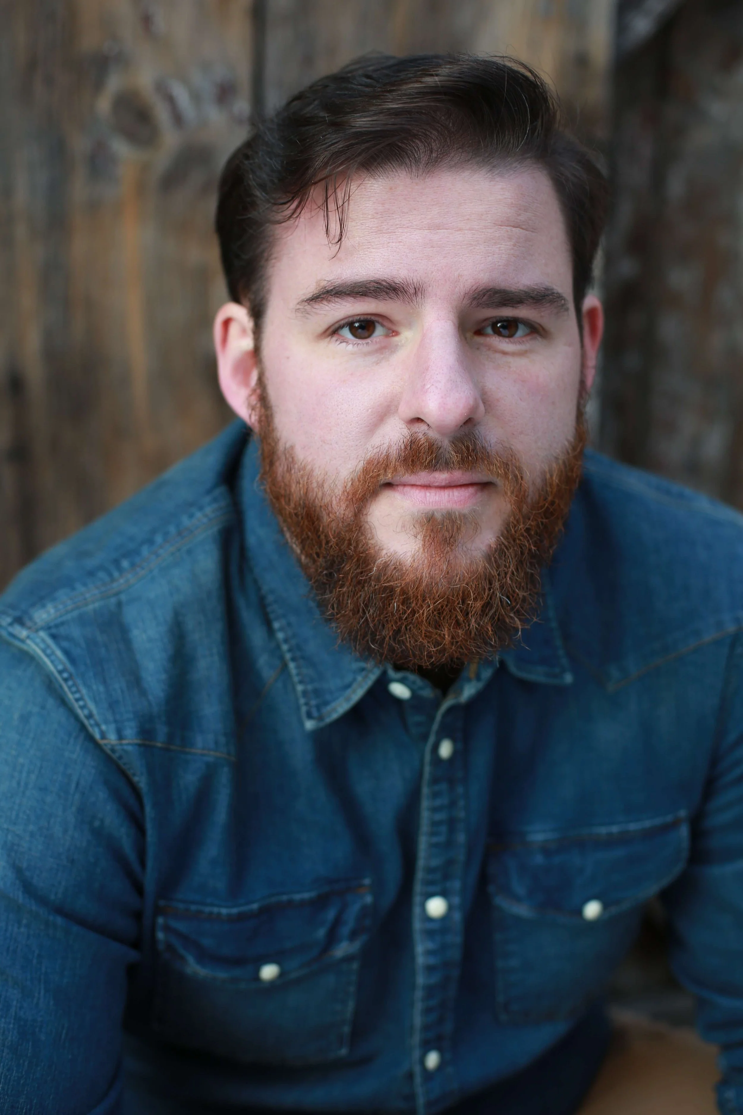 Heashot of Man with a reddish brown beard and brown hair looking at camera wearing denim button up