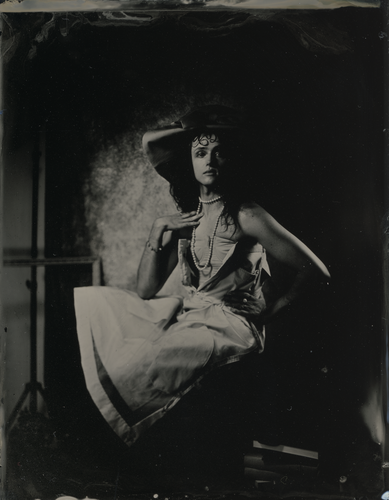 an editorial style photograph of a young man in high fashion wears on tintype, sitting in a photographic studio wearing a large hat