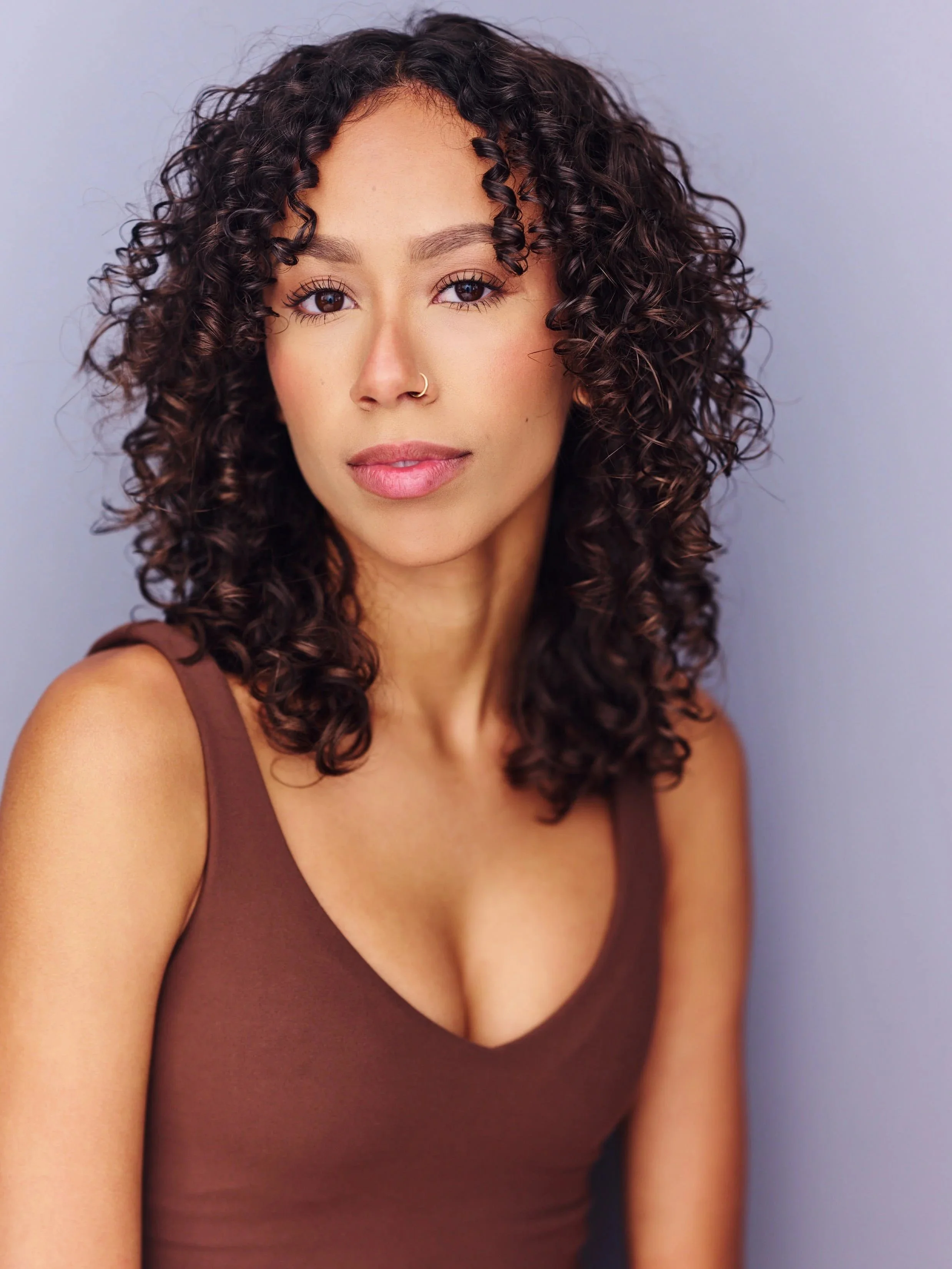 A woman with shoulder-length curly hair, wearing a brown sleeveless top and a nose ring, standing against a gray background.