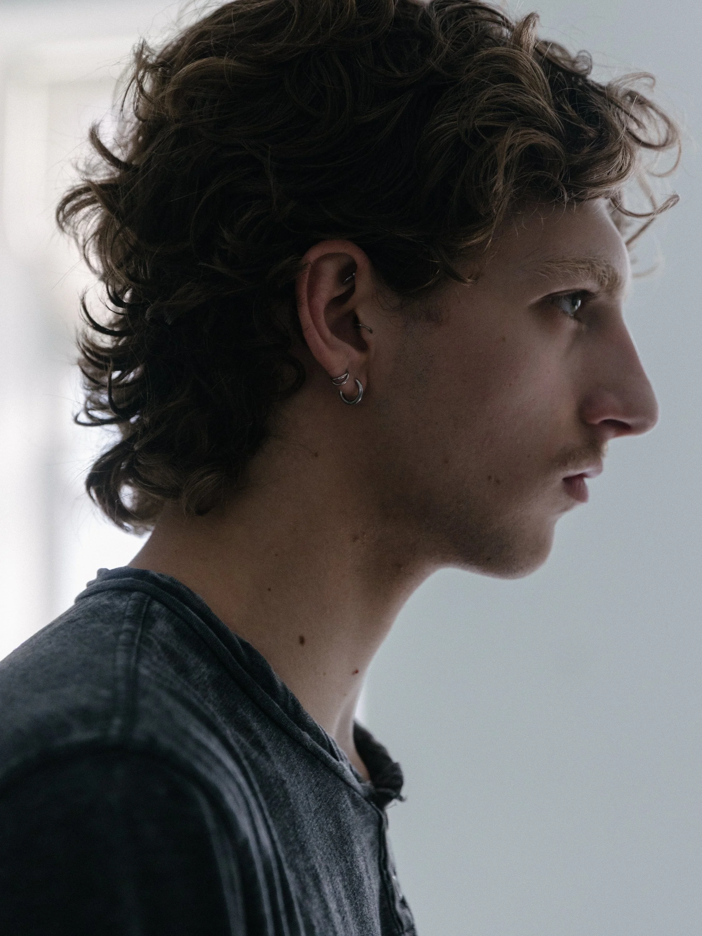 Close-up of a young person with short, curly brown hair and light skin, looking directly at the camera with a neutral expression, against a plain light background.