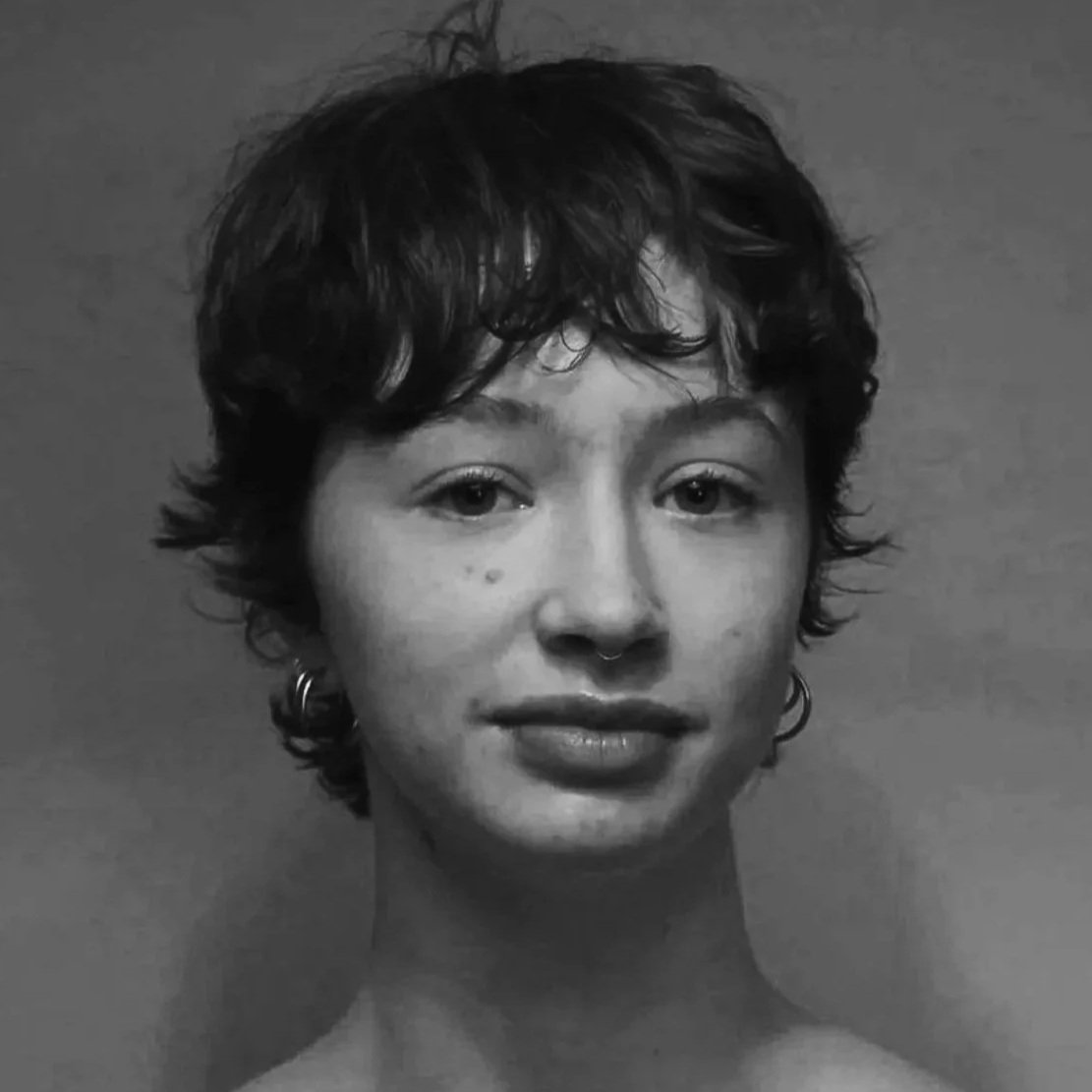 Black and white portrait of a young woman with short, curly hair, wearing earrings and a spaghetti strap top, looking directly at the camera.