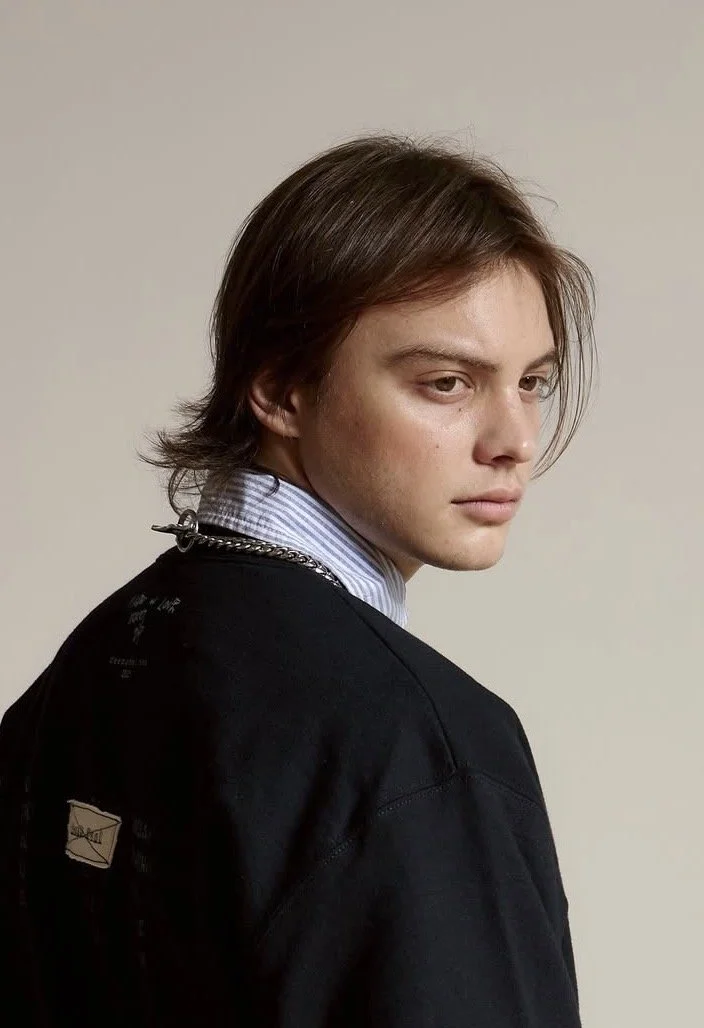 A young man with medium-length brown hair and fair skin, wearing a dark sweatshirt over a light blue collared shirt, looking over his shoulder against a plain beige background.