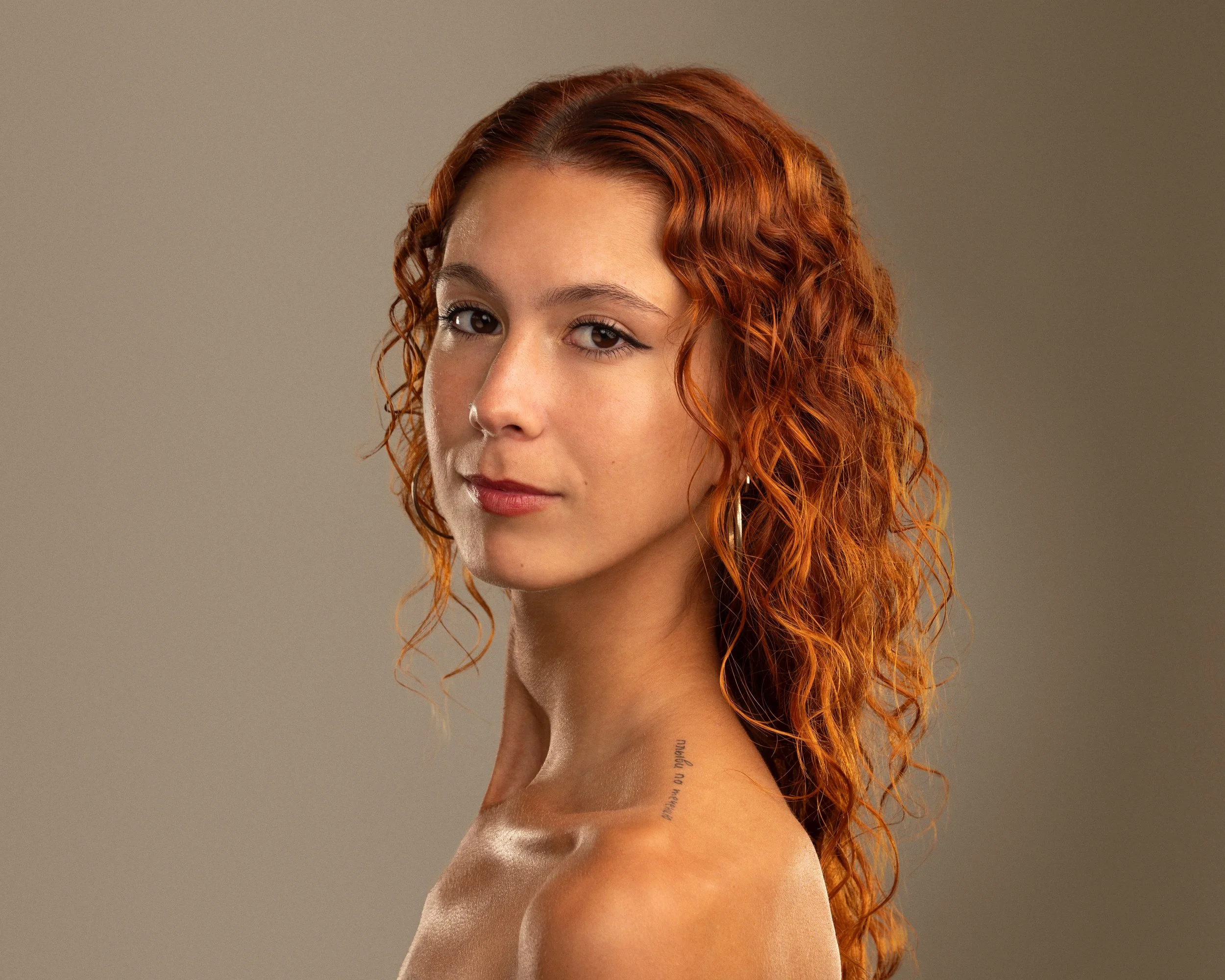A young woman with red curly hair and light skin, looking at the camera with a slight smile, showing a tattoo near her left shoulder on her neck.