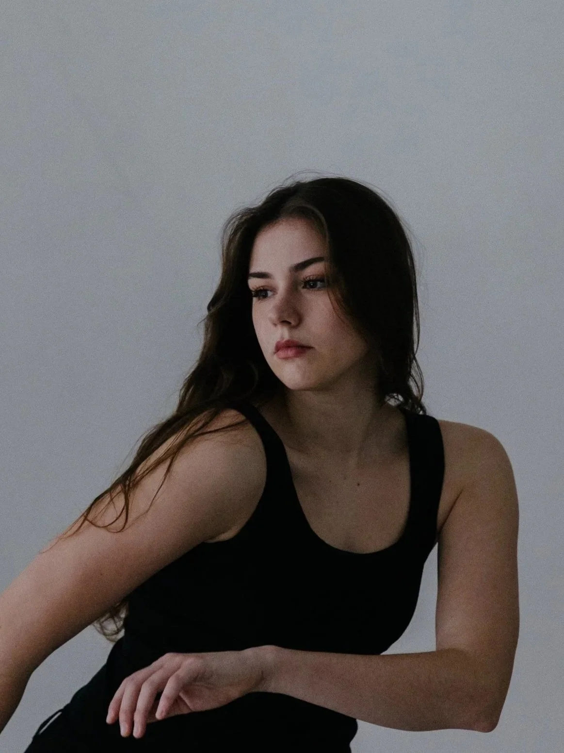 Portrait of a young woman with long brown hair and blue eyes, wearing a black top, against a gray background.