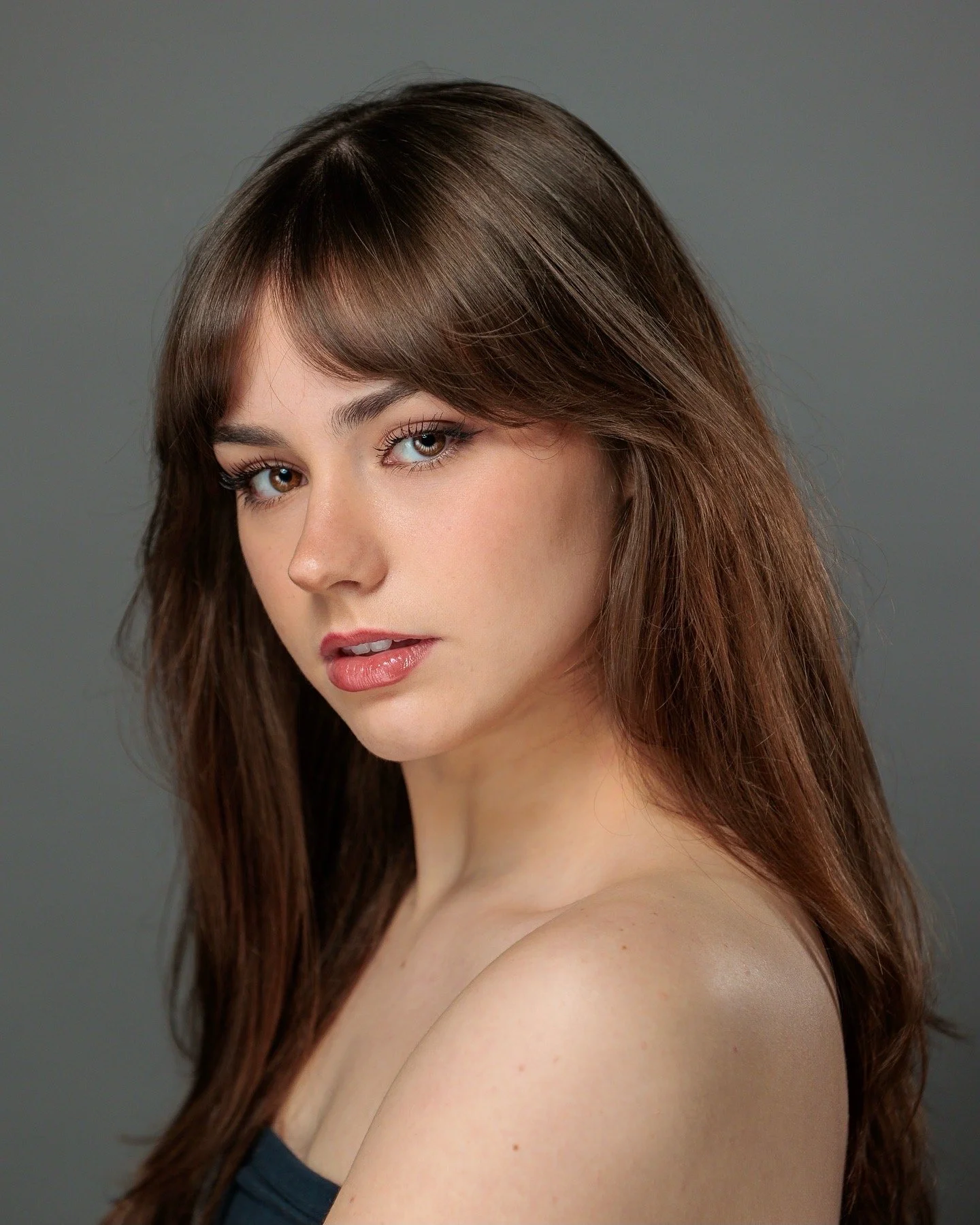 Portrait of a young woman with long brown hair and blue eyes, wearing a black top, against a gray background.