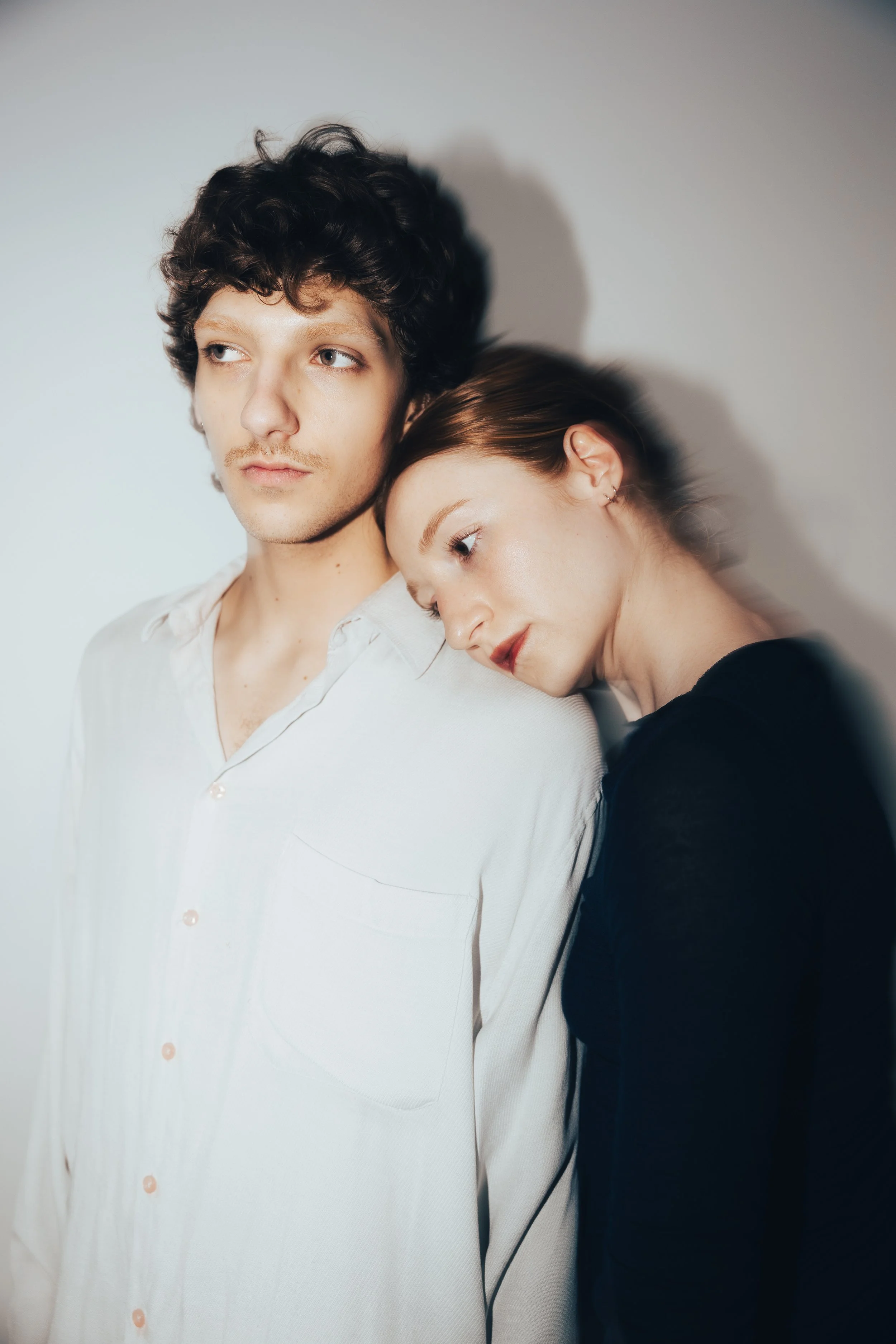 A young man with curly dark hair and a light mustache, wearing a white shirt, stands next to a young woman with reddish brown hair, who is resting her head on his shoulder, wearing a black top. They are against a light gray background.
