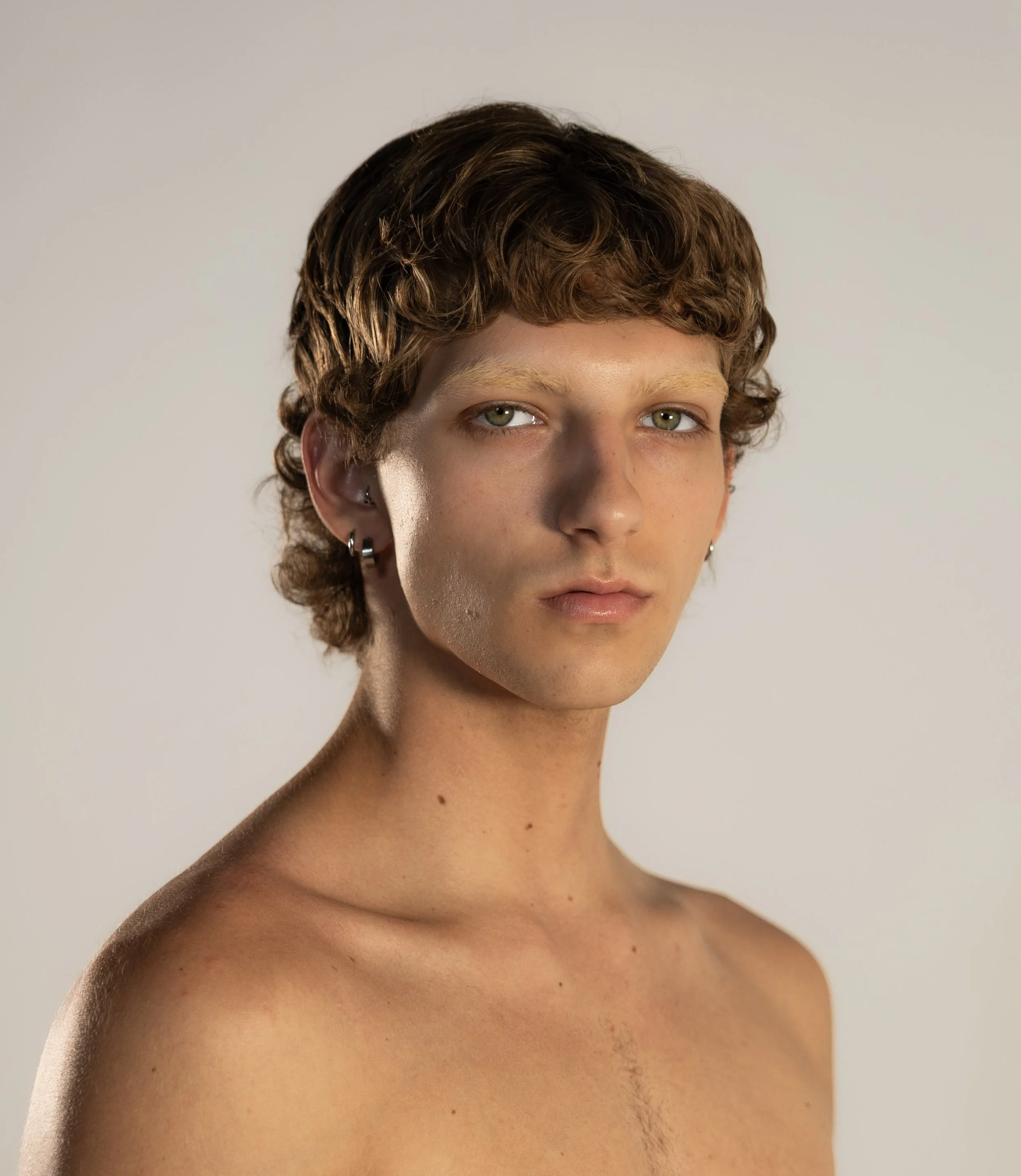 A young person with short, curly brown hair, light skin, and piercing blue eyes, looking directly at the camera against a plain neutral background.