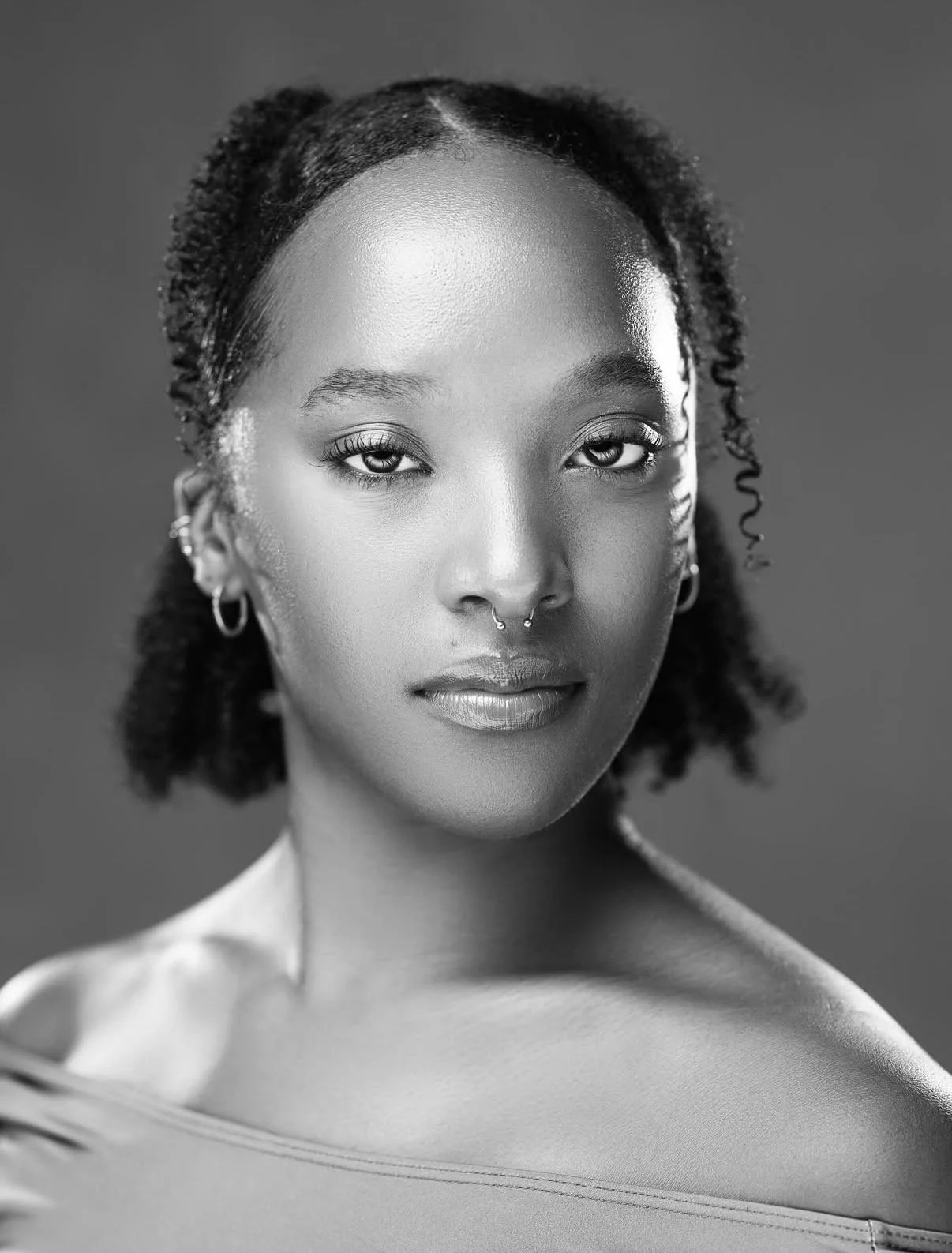 Black and white portrait of a young woman with natural curly hair, pierced nose, hoop earrings, and wearing an off-shoulder top.