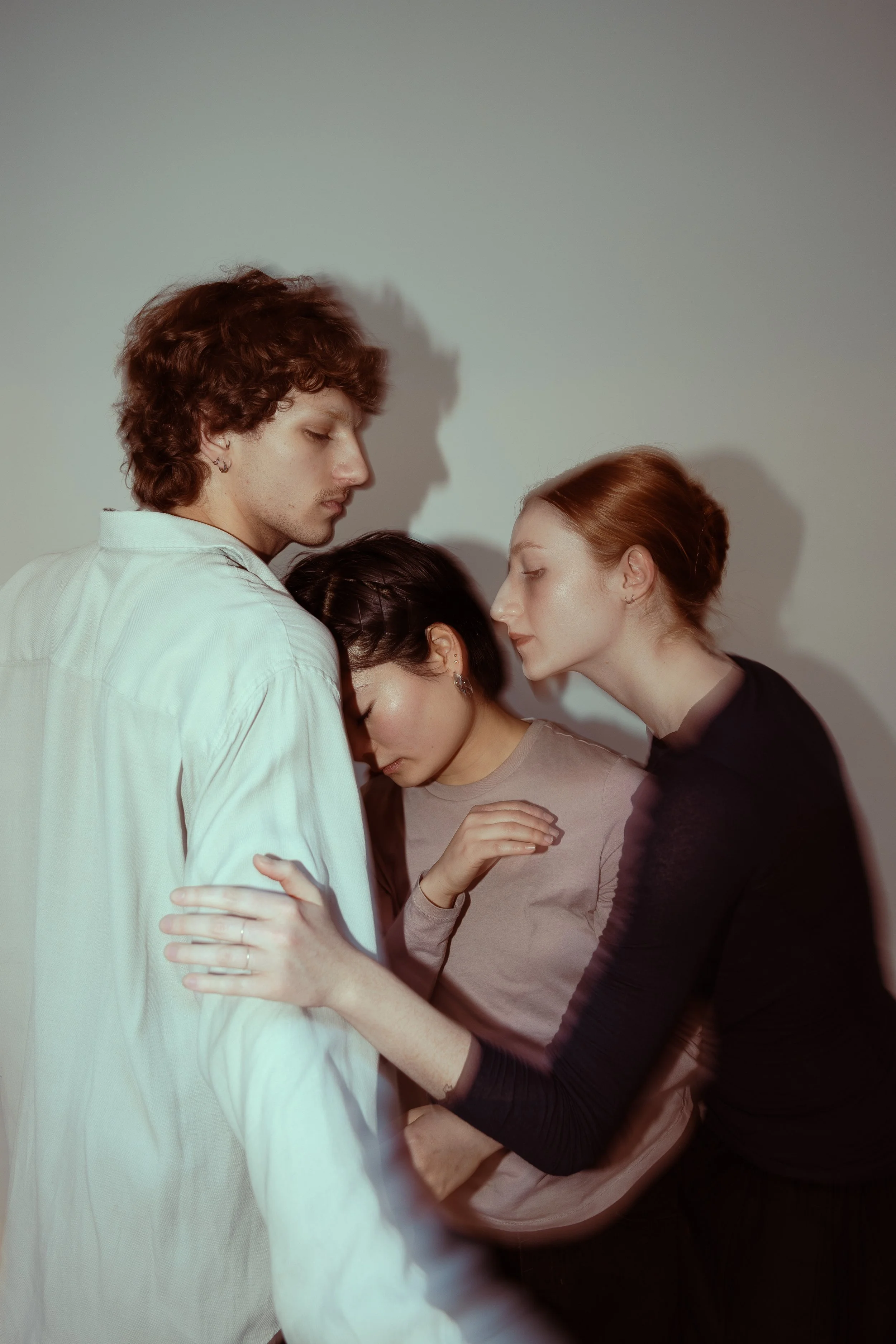Three people closely embracing, their eyes closed, in an intimate moment against a plain background.