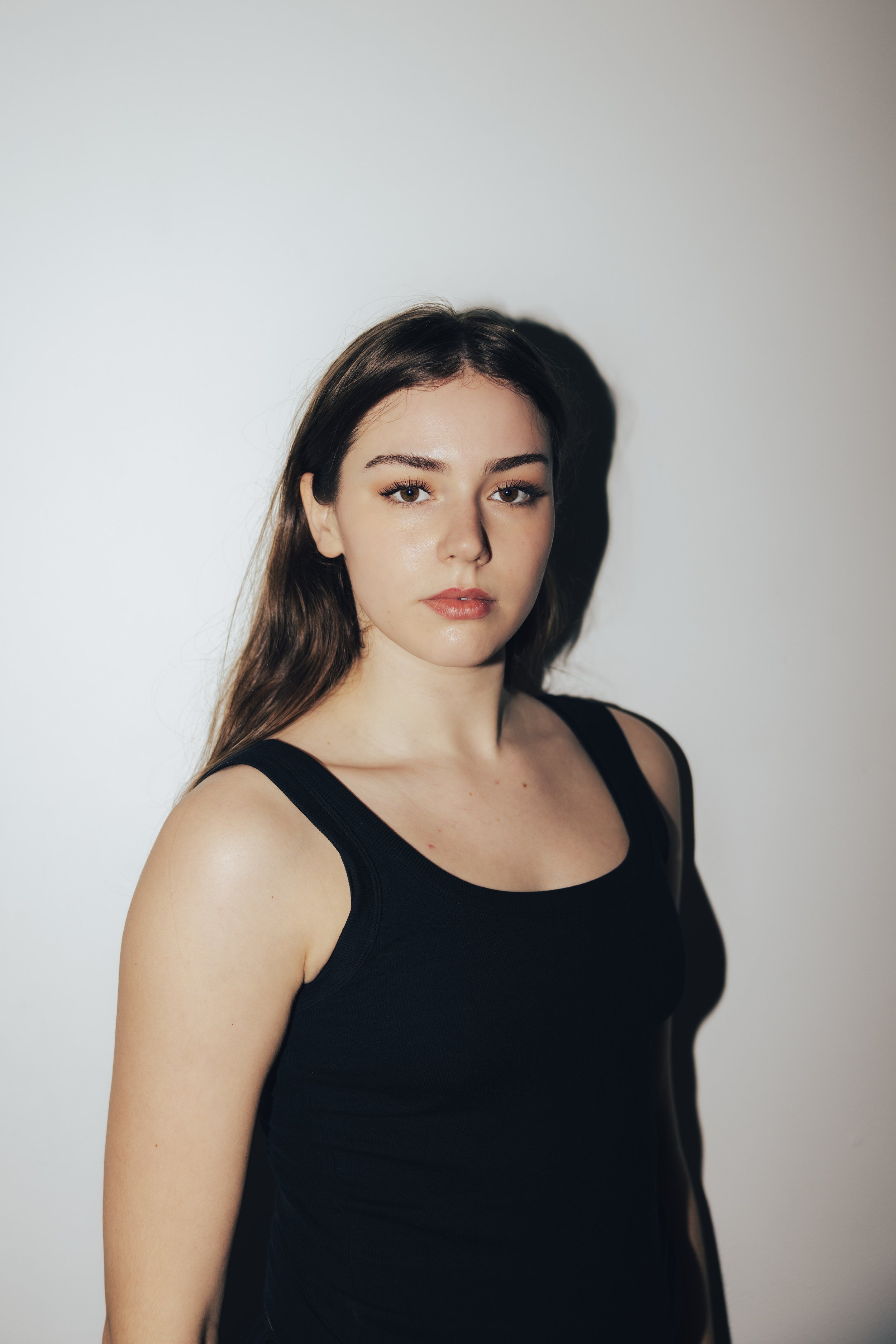 Portrait of a young woman with long brown hair and blue eyes, wearing a black top, against a gray background.