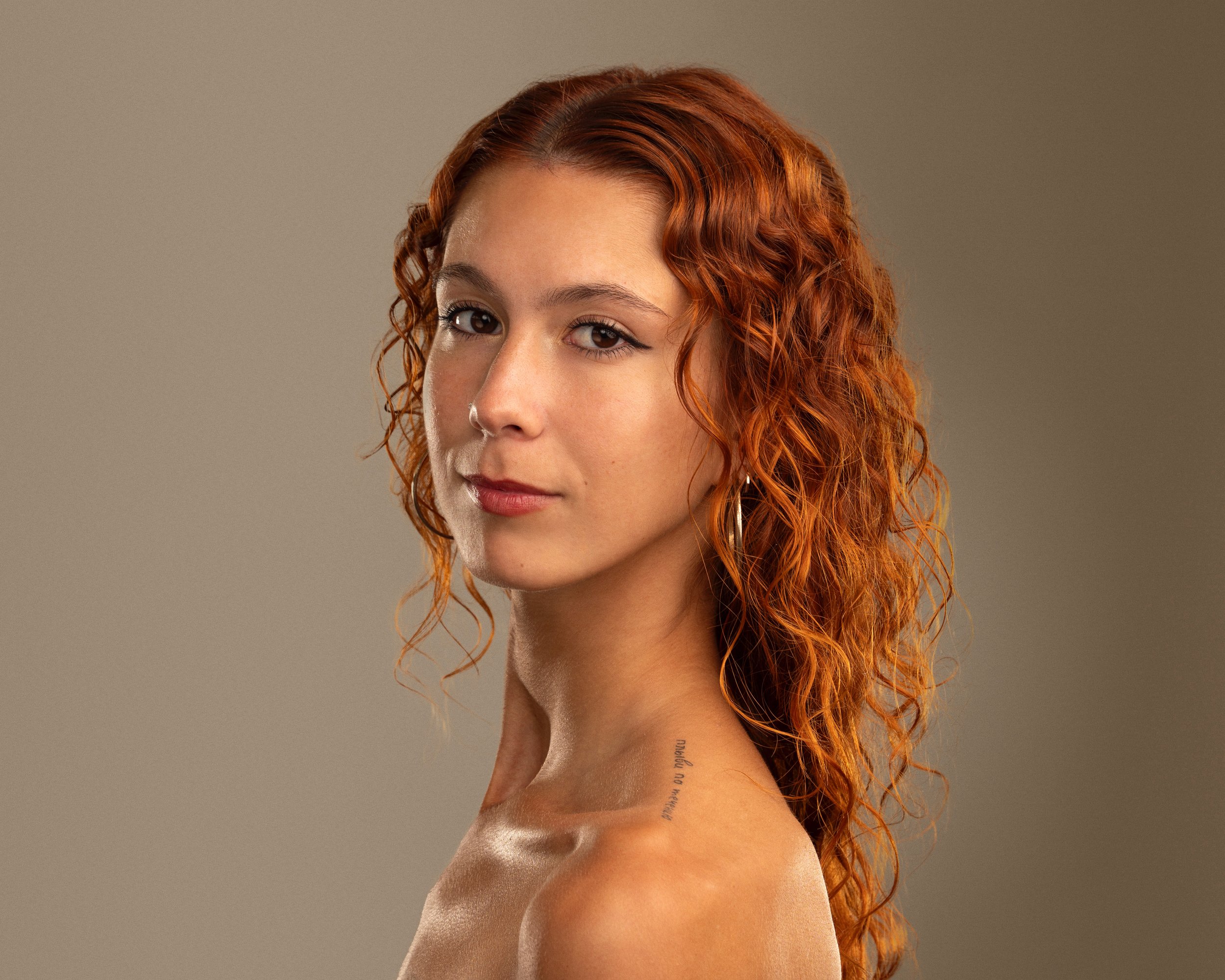 A woman with wavy red hair, light skin, and a tattoo on her shoulder, looking at the camera against a neutral background.