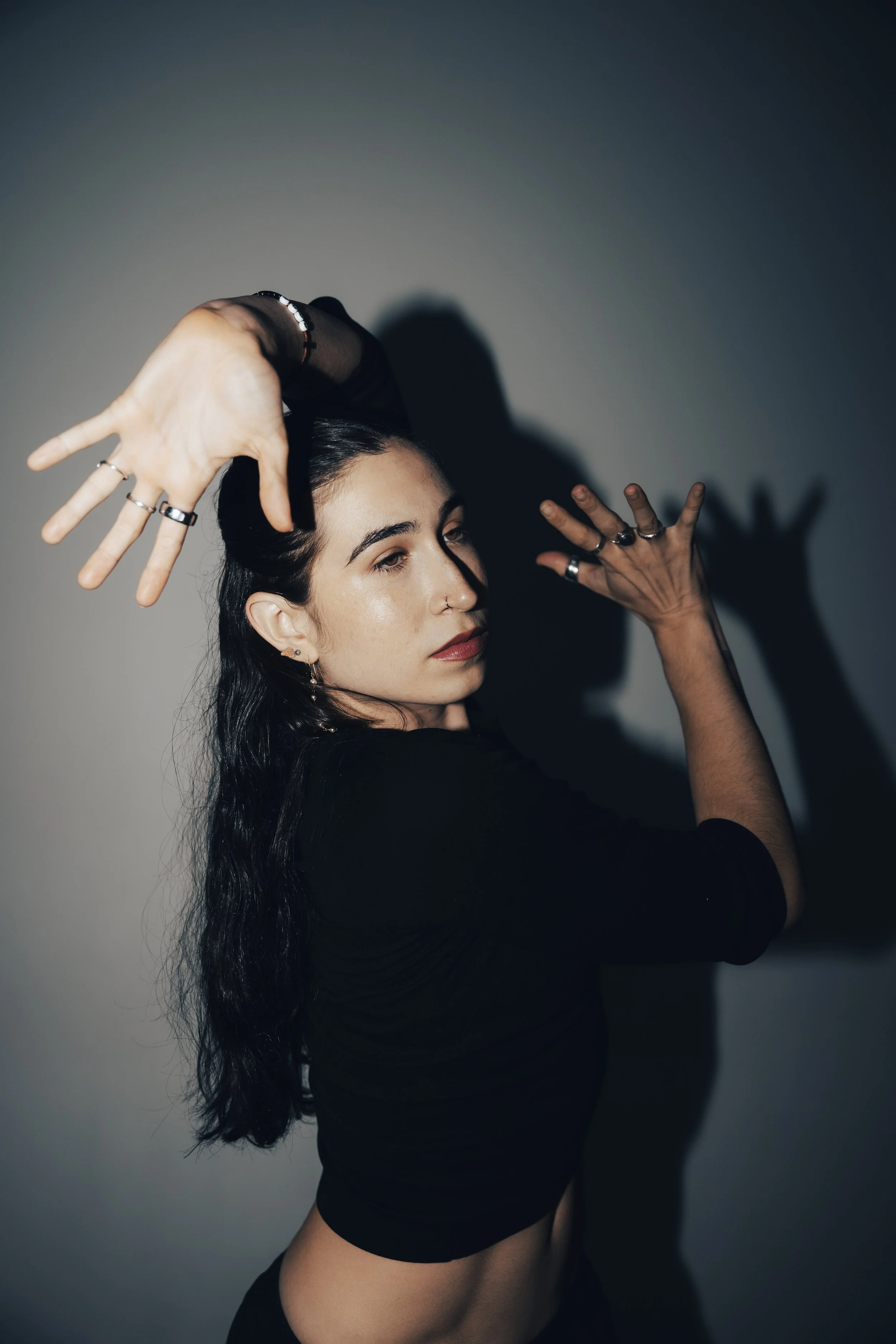 A woman with long dark hair and earrings, wearing a black top, is posing against a light-colored wall with her arm outstretched. There is a motion blur effect in the image.