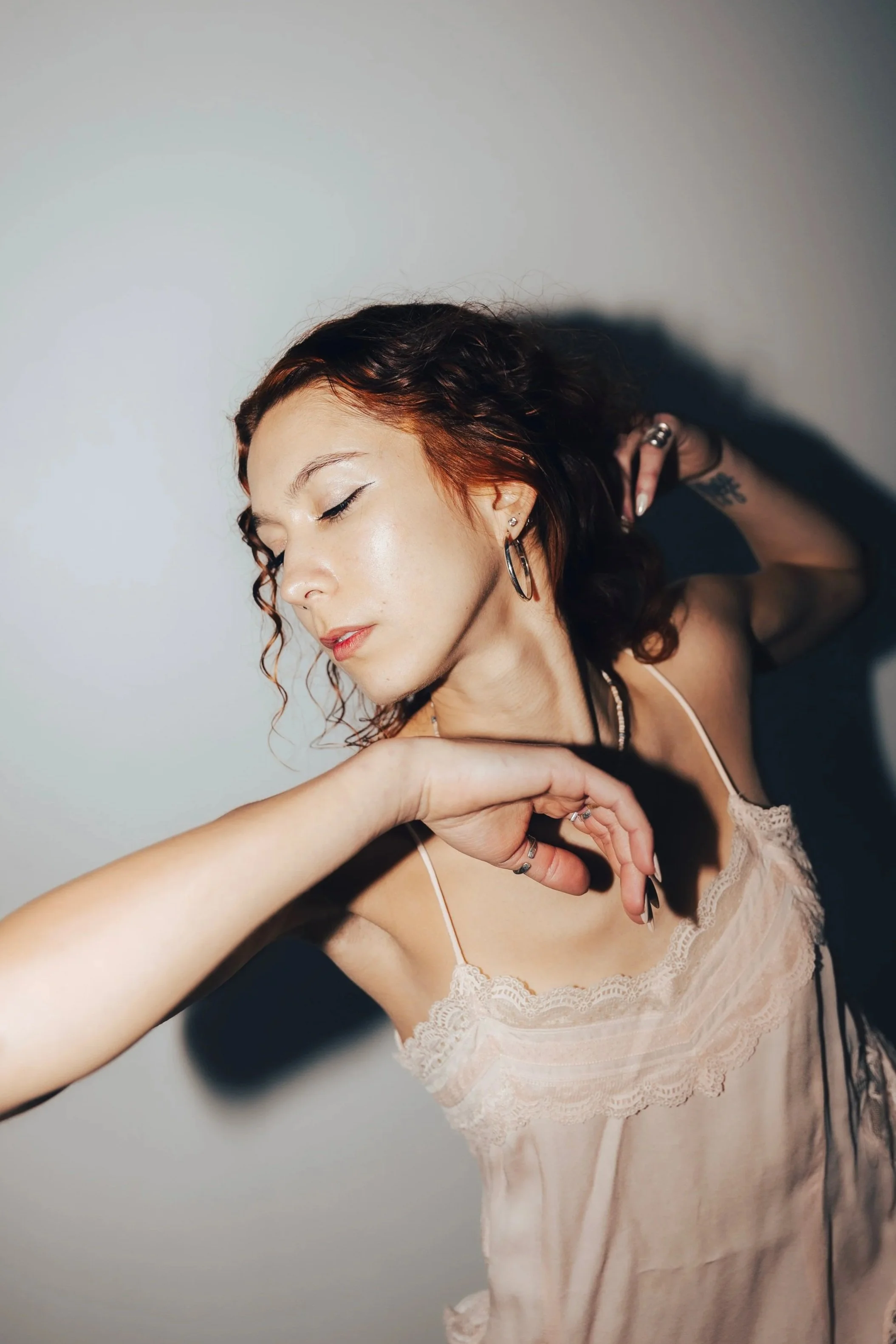 A young woman with red curly hair and light skin, looking at the camera with a slight smile, showing a tattoo near her left shoulder on her neck.