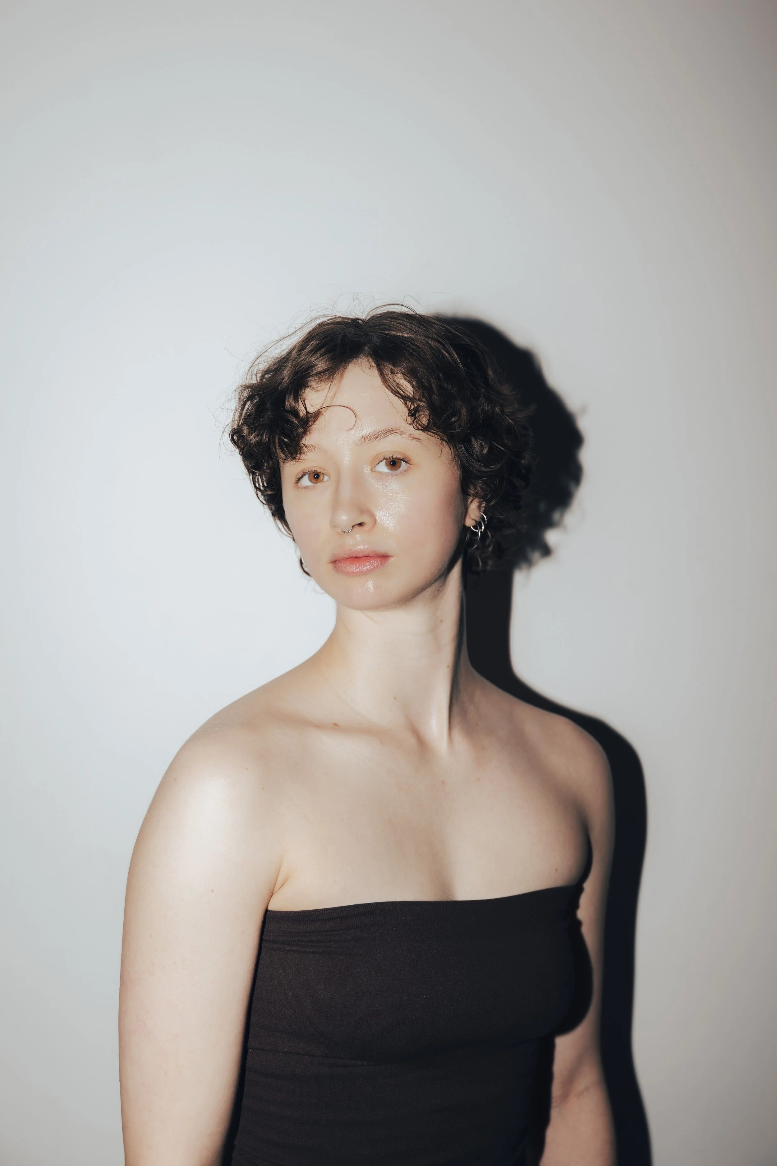 Black and white portrait of a young woman with short, curly hair, wearing earrings and a spaghetti strap top, looking directly at the camera.