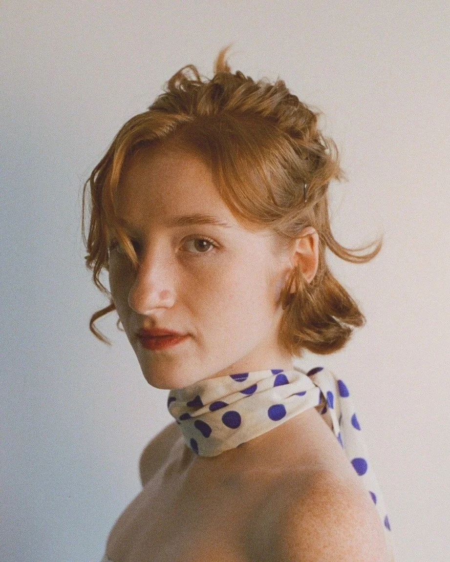 Portrait of a young woman with short wavy red hair, looking over her shoulder, wearing a polka dot scarf around her neck, against a plain background.