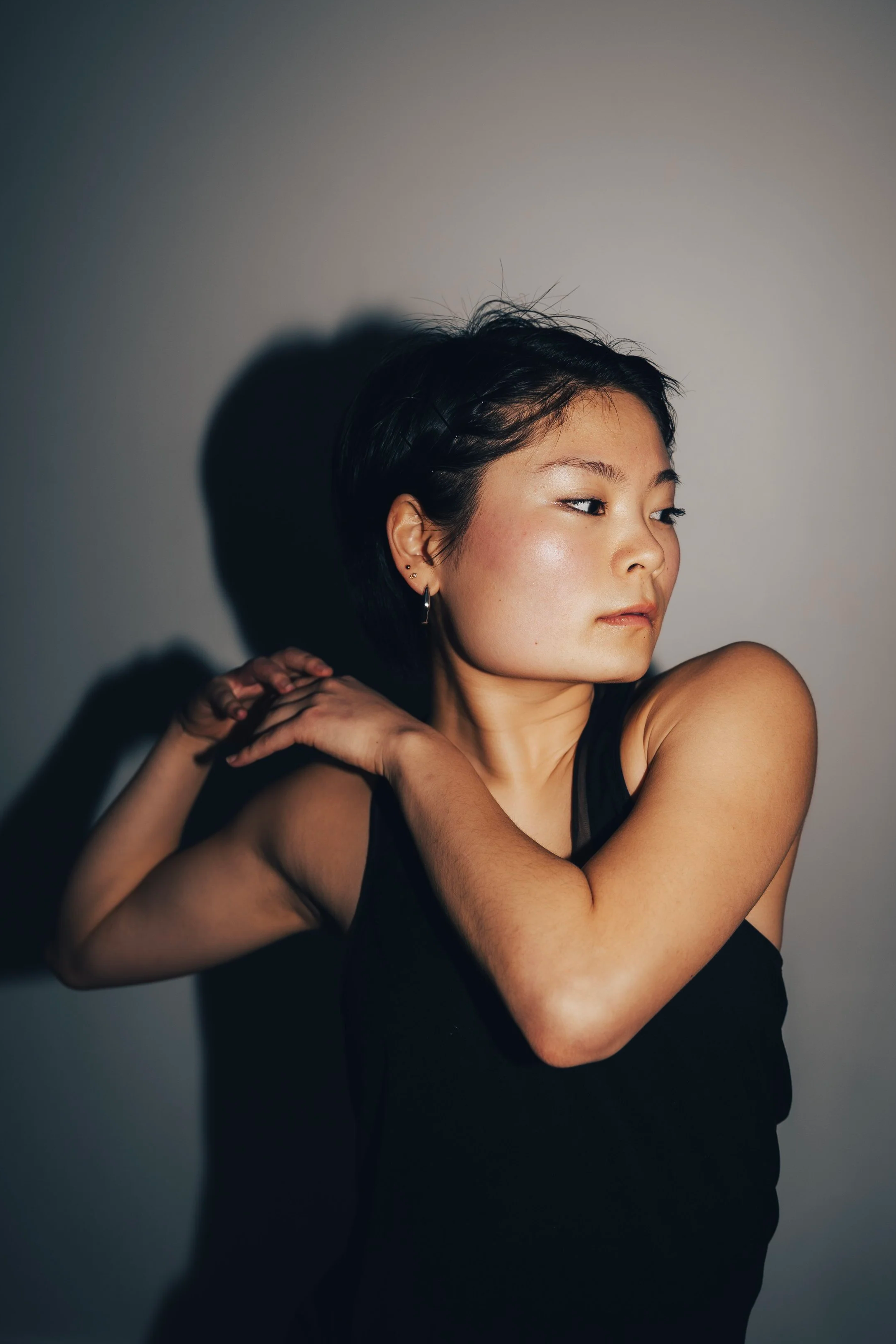 Portrait of a young woman with short black hair, wearing a black lace spaghetti strap top, looking confidently at the camera with a slight smile, against a plain light gray background.