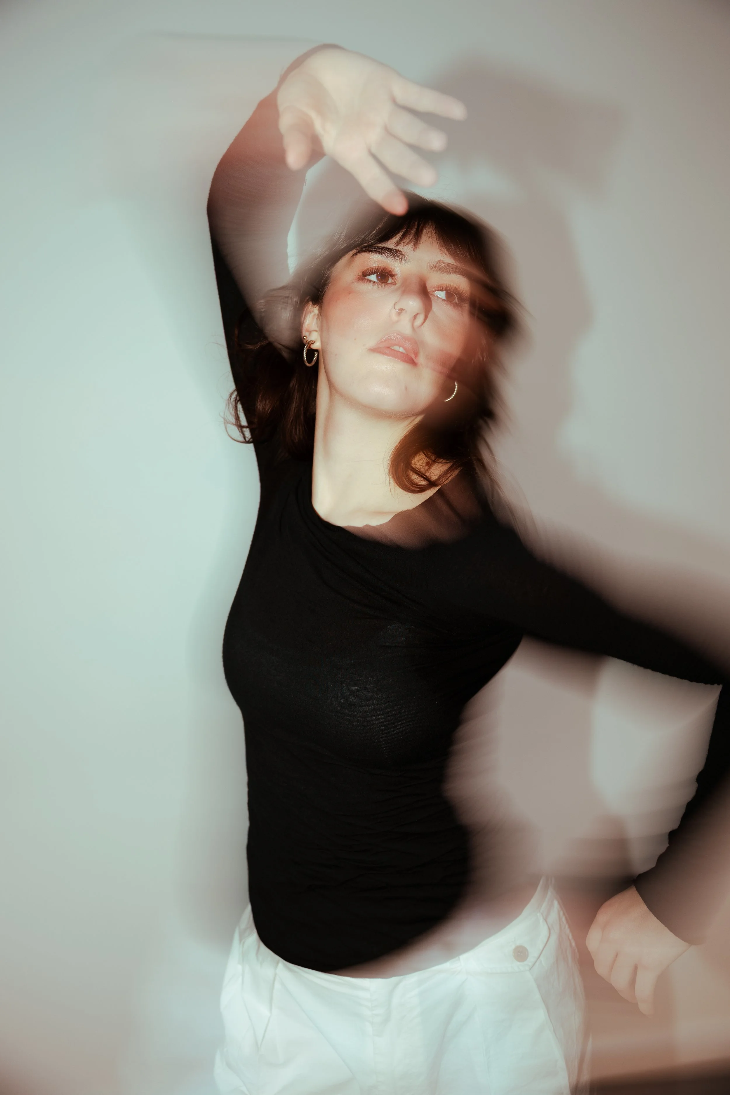 A young woman with shoulder-length brown hair and hoop earrings poses against a plain wall. She is stretching her left arm above her head, creating a blurred motion effect that obscures part of her face and body. She is wearing a black long-sleeved top and white pants.