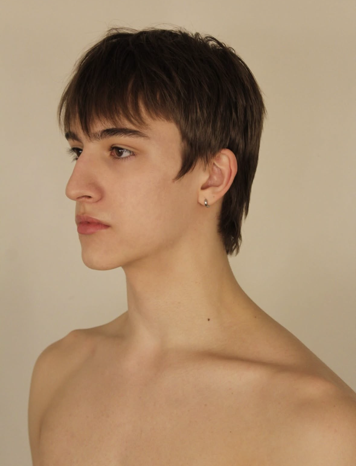 Side profile of a young man with short brown hair, wearing a small hoop earring, against a plain beige background.