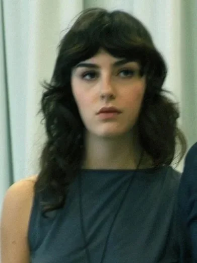 A woman with shoulder-length dark brown hair standing in front of white curtains.