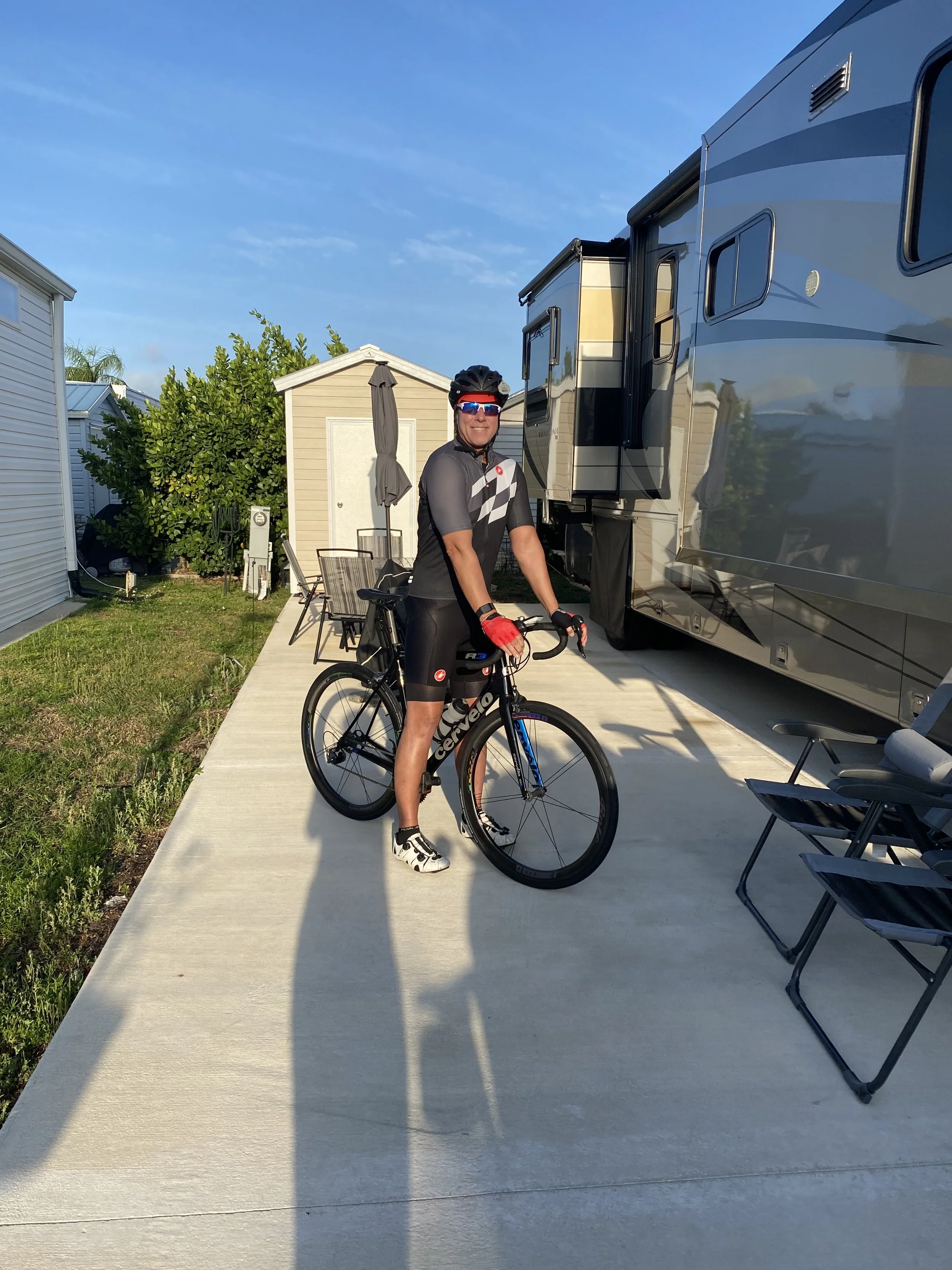 A man in cycling attire with a helmet and sunglasses standing next to a black and blue road bicycle on a concrete patio near a large RV, with outdoor furniture and a small shed in the background on a sunny day.