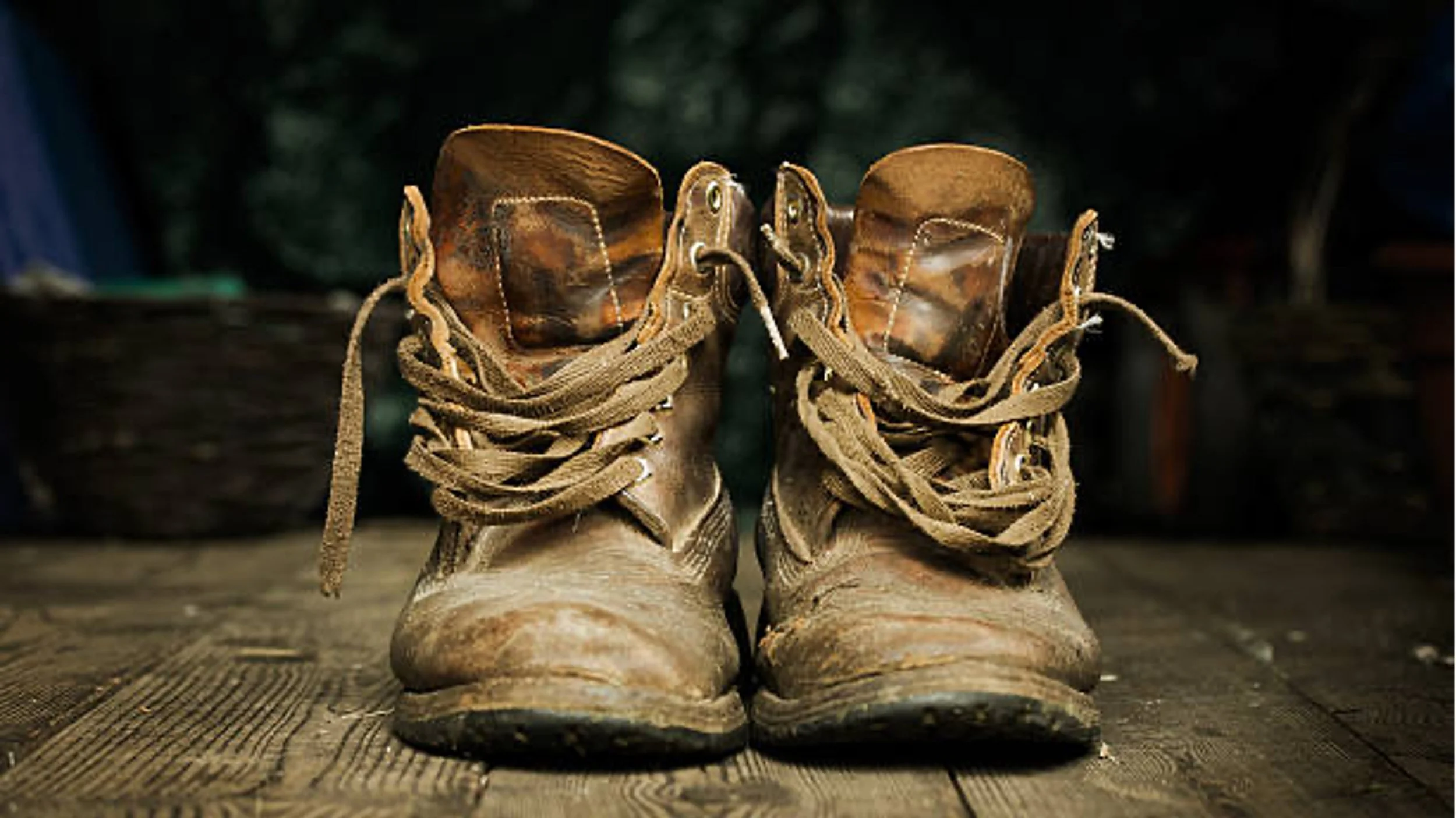 A pair of well-worn, brown leather work boots with laces on a wooden surface, with a blurred background of trees and outdoor scenery.