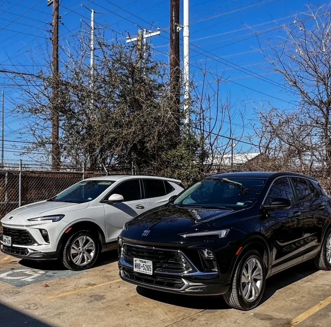 Two new cars, a white SUV and a black SUV, parked in a parking lot with a chain-link fence, trees, and utility poles in the background under a clear blue sky.