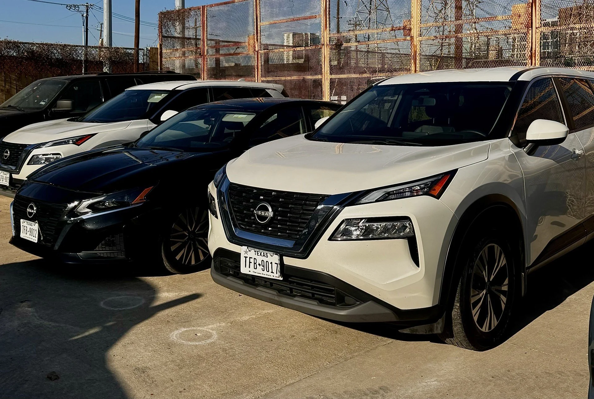 A row of white, black, and dark-colored Nissan vehicles parked in a lot, with a chain-link fence and industrial buildings in the background.