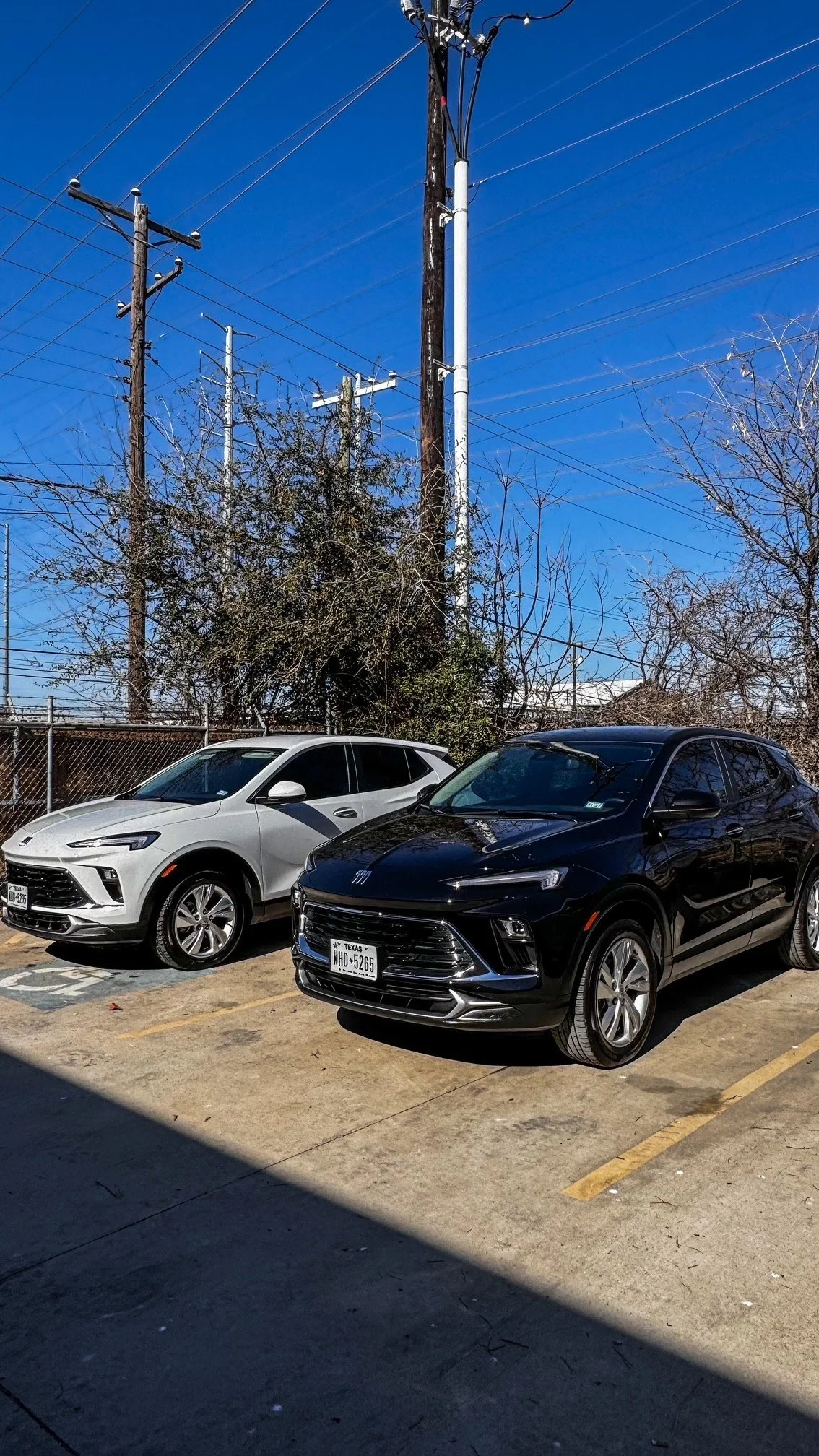 Two cars parked in a parking lot, a white SUV and a black SUV, with utility poles and wires in the background, under a clear blue sky.