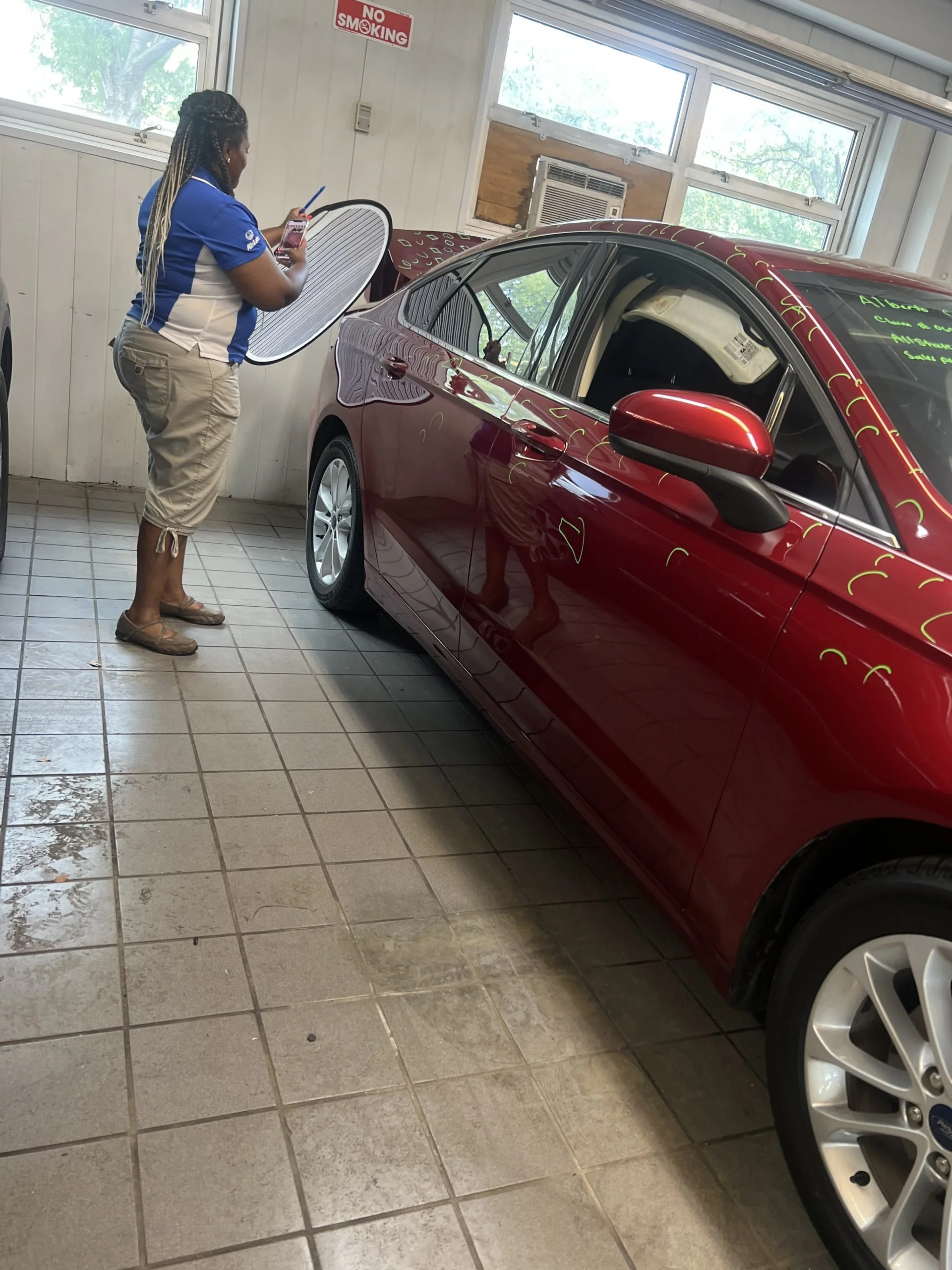 A woman in a blue and white polo shirt standing inside a car dealership, holding a tablet, inspecting a red car with writing on the windshield, tiled floor, windows in the background, and an air conditioning unit. There is a 'No Smoking' sign on the 