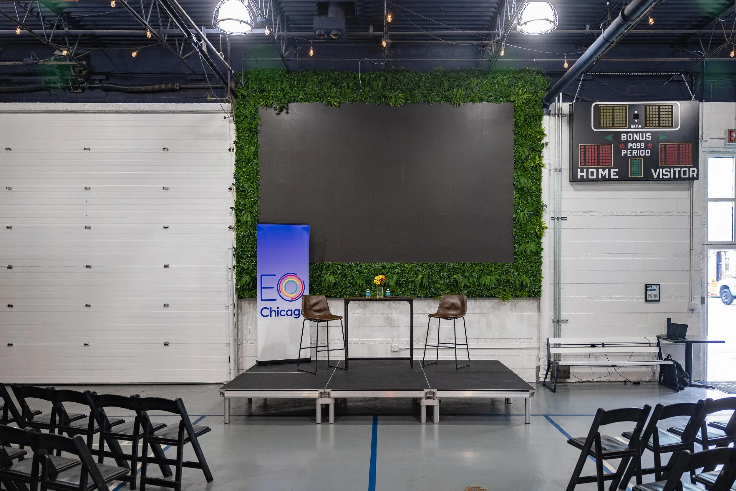 An indoor event space with black walls and ceiling, decorated with green plants around a large blank black display screen. There are chairs arranged facing the stage, and a small stage with two chairs and a table in front of the screen. To the right,