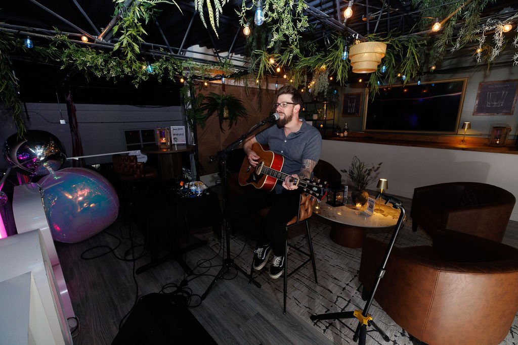 A man with glasses and a beard playing an acoustic guitar and singing into a microphone in a cozy, dimly-lit venue decorated with hanging plants and string lights.