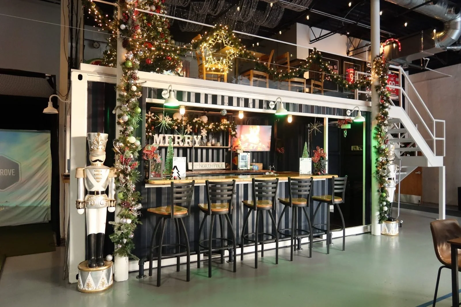 Christmas-themed bar area decorated with garlands, ornaments, and lights, featuring a Nutcracker soldier statue in front, a shelf with holiday decorations, and a staircase with garland on the right side.