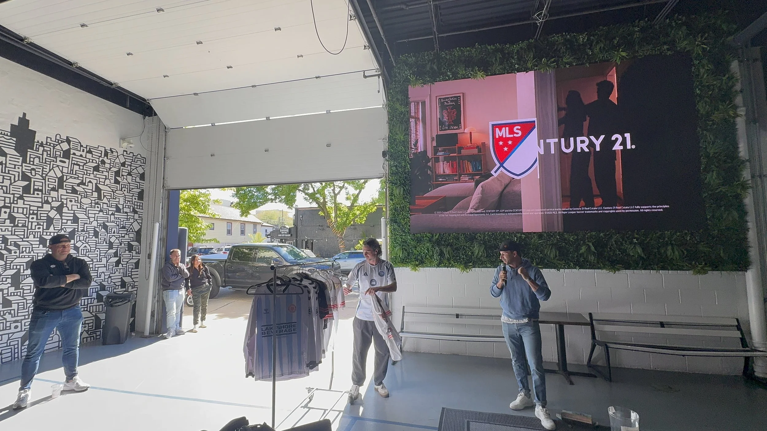 Indoor gathering with promotional merchandise, including shirts, with a large digital banner displaying MLS Century 21 logo, people talking and standing near a white brick wall, with some standing outside visible through an open door.